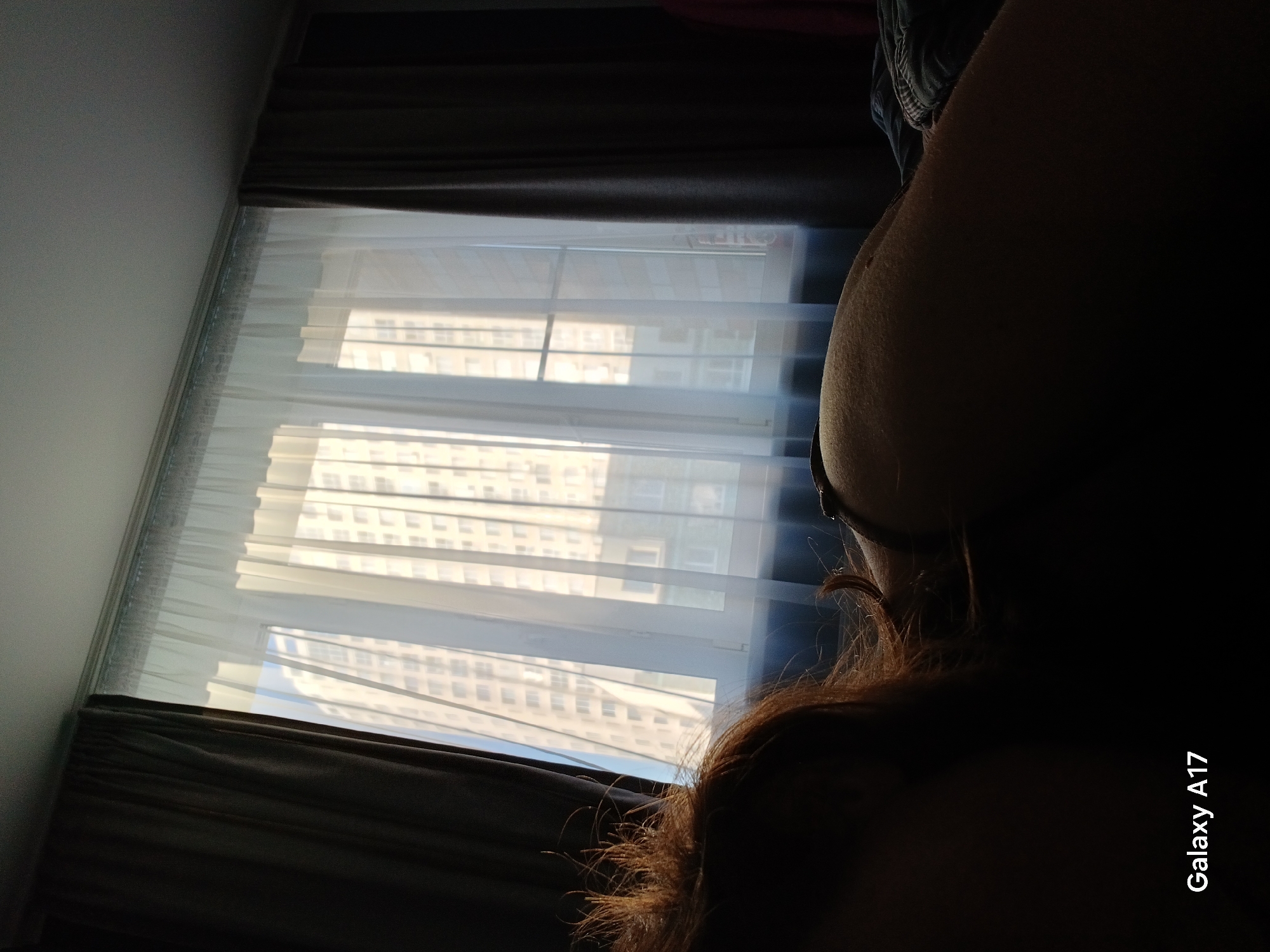 An interior view through a window with white blinds and brown curtains reveals a building facade in Yekaterinburg, Russia. The foreground is dark, obscuring details of a person with brown hair, except for the arm. The photo includes a "Galaxy A17" watermark.