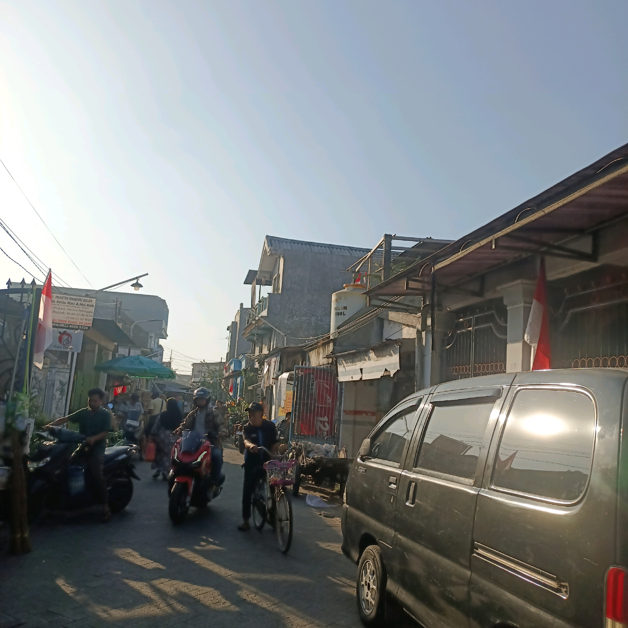 The image depicts a street scene in Surabaya, Indonesia, on a clear, bright day, likely in the late afternoon given the long shadows. The street is paved and appears to be moderately busy with both motorized and pedestrian traffic.
In the foreground, a dark-colored van is parked on the right side of the street, its surface reflecting the bright sunlight. Further down the street, several motorcycles are visible. On the left side, a man in a green shirt is riding a dark motorcycle. Next to him, another person is on a red motorcycle. A person is also seen walking a bicycle with a basket in the middle of the street.
On the left side of the street, buildings line the road. One building has a sign that reads "PRAKTEK MANDIRI BIDAN" and lists a name and contact information. A red and white Indonesian flag is prominently displayed on a pole in front of this building. Other buildings appear to be shops or residences, some with awnings and various signs. A green umbrella is visible over a stall or shop.
On the right side of the street, more buildings are visible, some appearing to be multi-story. A large white water tank with "PROF TANK 1100 L" written on it is mounted on one of the buildings. Another Indonesian flag is displayed on a building further down the street. The overall impression is of a bustling urban environment.