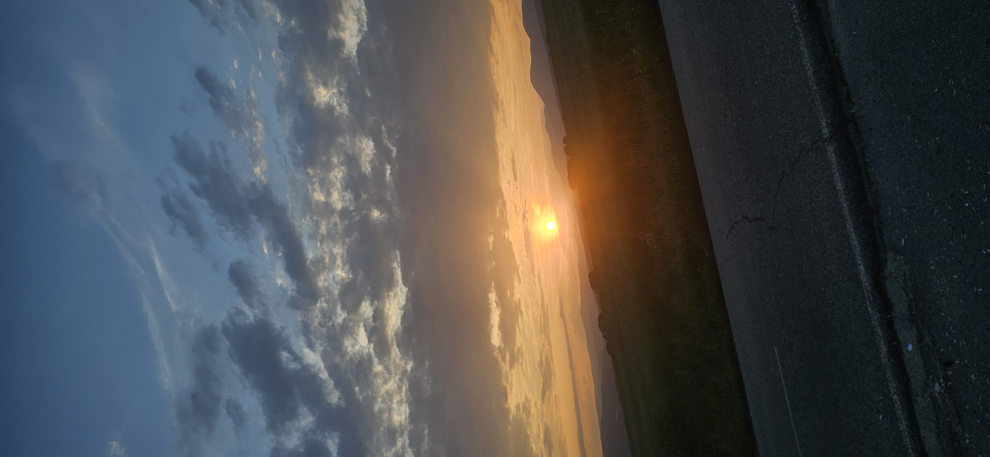 The image captures a sunset over a rural landscape in Momin Prohod, Bulgaria. The sun, appearing as a bright, intensely glowing golden orb, is positioned low above the horizon, slightly to the left of the vertical center. Its light illuminates the sky directly above the horizon with strong orange and yellow hues. Above this, a layer of dark grey altocumulus clouds with patches of bright orange and white highlights extends across the middle of the sky. The uppermost portion of the sky is a clearer, lighter blue with a few faint, wispy clouds. The mid-ground reveals rolling green fields and distant mountain ranges, which are partially silhouetted against the bright sky and partially illuminated by the warm light. In the foreground, the edge of a dark asphalt road with a faded white shoulder line and several visible cracks traverses the bottom portion of the frame, angled upwards from the lower left towards the lower right. No individuals, animals, or specific man-made structures are discernible within the scene beyond the road.