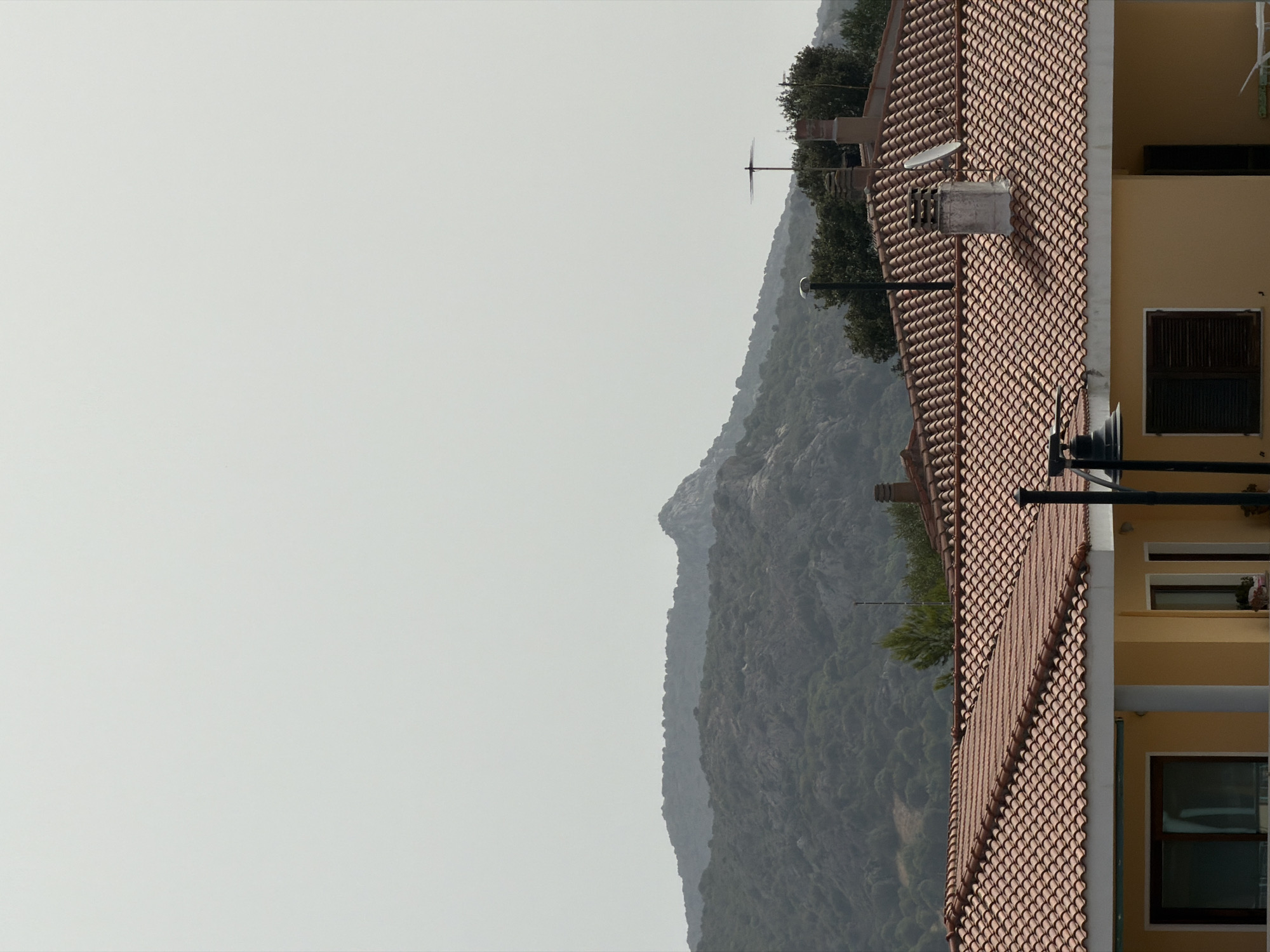 The image shows a residential building with a tiled roof, characteristic of Olbia, Italy. The building is painted yellow and has dark-shuttered windows and white columns. In the background, there are hills covered with trees and rocks, with a clear sky above. The overall scene depicts a quiet, rural or semi-rural setting, possibly on the outskirts of the city. The architecture of the building suggests a typical Mediterranean style. The composition of the image emphasizes the contrast between the man-made structure and the natural landscape.