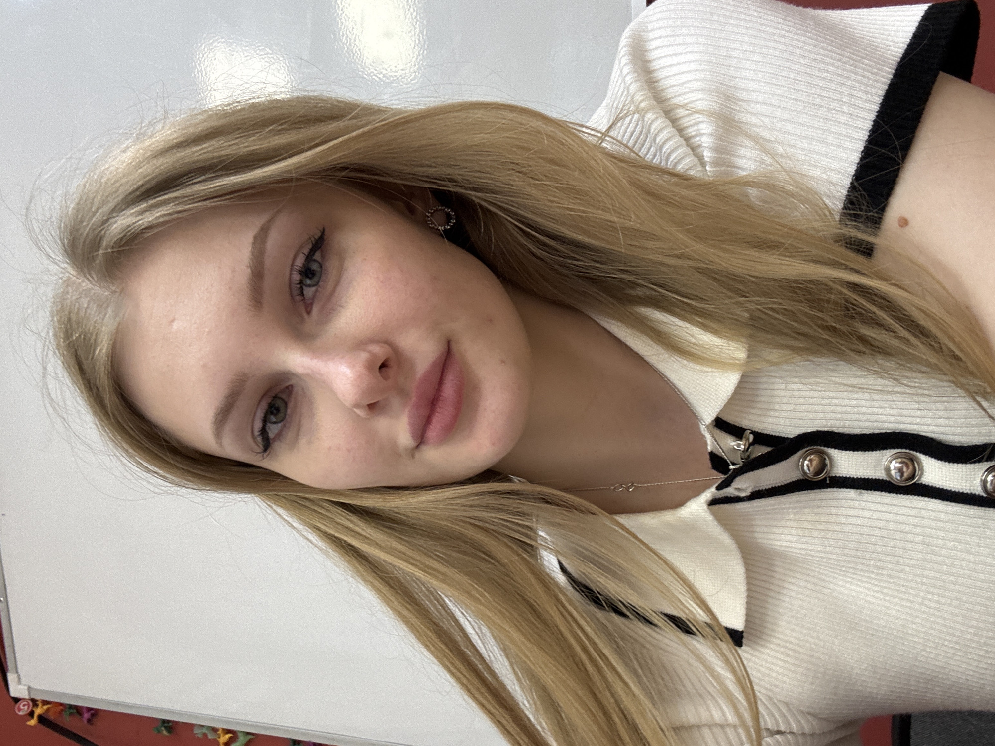 A young woman with blonde hair is captured in a close-up, slightly angled portrait. She is wearing a white, collared top with black trim and silver buttons. Her expression is soft and she is wearing earrings. The background appears to be a white surface, possibly a wall. The location is Perm, Russia.