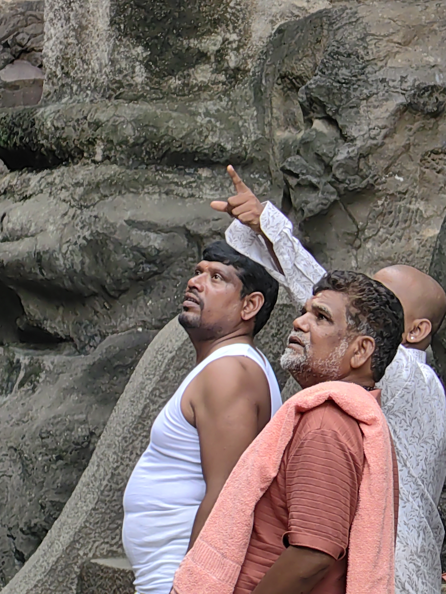Three men are standing in front of a carved rock formation in Ellora, India. The man on the left is wearing a white tank top. The man in the center is wearing a rust-colored shirt and has a pink towel draped over his shoulder. The man on the right, who is mostly obscured, is wearing a white patterned shirt and has a bald head. One of the men is pointing upwards, seemingly at the rock formation. The rock formation appears to be ancient and weathered.