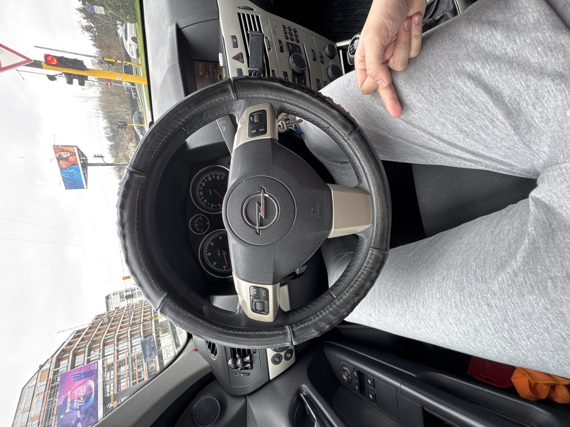 A view from inside a car with an Opel steering wheel. Outside the car, there is a red traffic light, a building with construction cranes, and a billboard advertising "Iqos Iluma Electric." The car is presumably in Sofia, Bulgaria, based on the Cyrillic text on the billboard. A person wearing gray sweatpants is visible in the passenger seat, pointing down with their index finger. Keys are hanging from the steering wheel. The dashboard and interior of the car are visible.