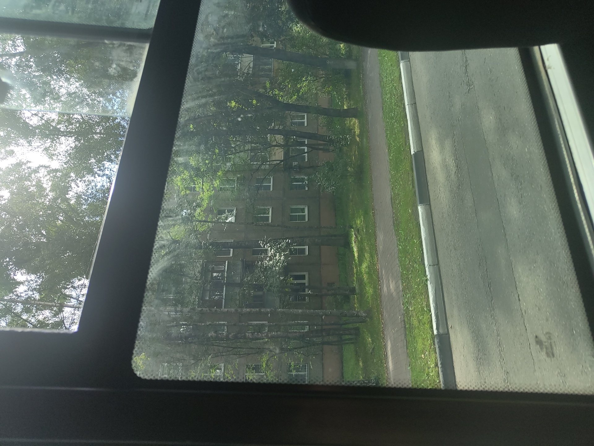 The image depicts a view through a window of a building in Ramenskoye, Russia. The building is a multi-story apartment block, characterized by its grey facade and numerous windows with white frames. Trees with green foliage are interspersed in front of the building, casting shadows and creating a somewhat obscured view. A paved sidewalk runs in front of the building, bordered by a grassy area and a road with a curb. The sun appears to be shining, as indicated by the bright sky and dappled light on the trees and ground. The perspective suggests the photo was taken from a moving vehicle or a window in another building. The scene is a typical urban residential environment.