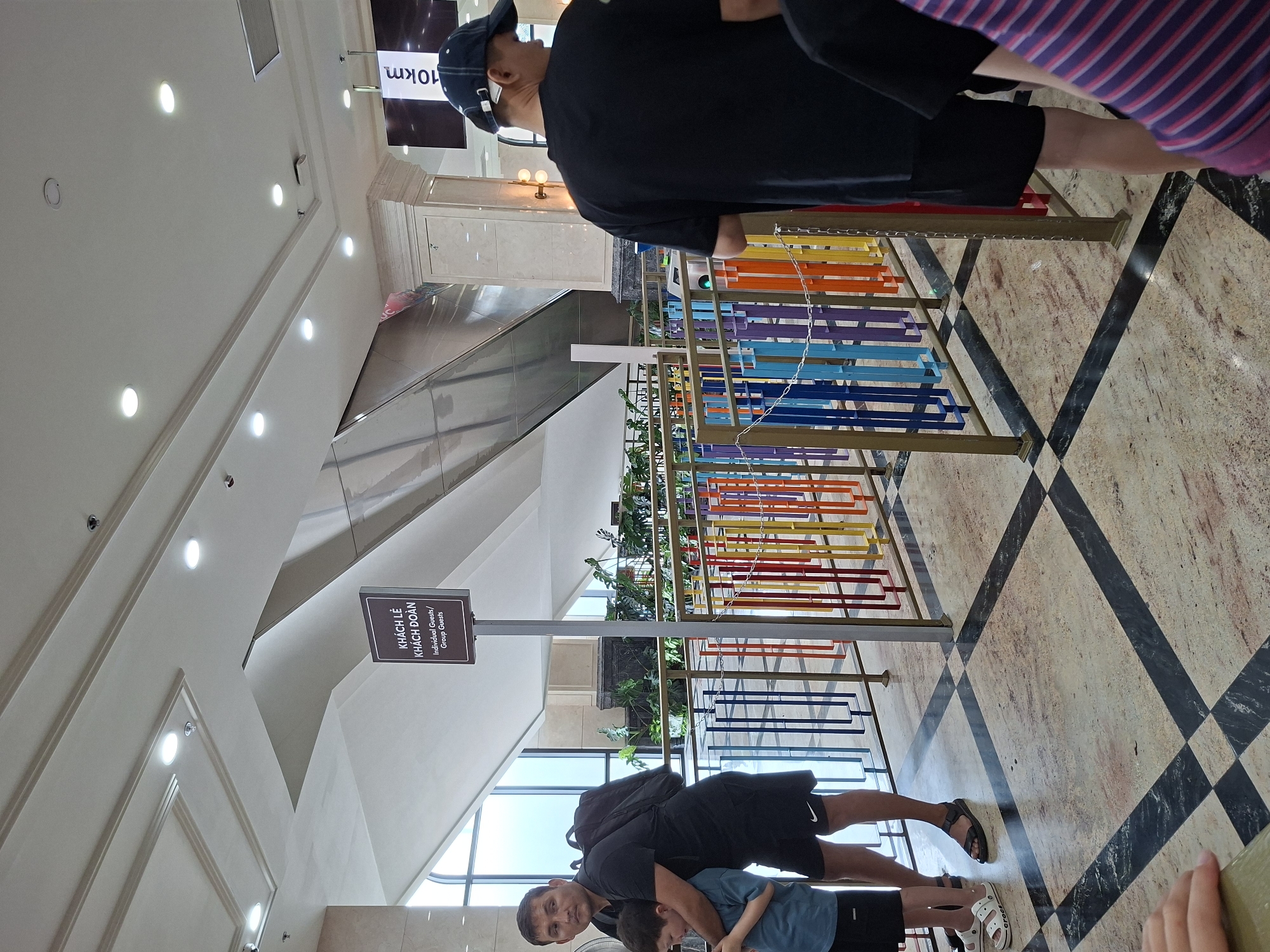 Here is a description of the image:
**Scene:** The image depicts the interior of a building, likely a hotel or shopping center, in Nha Trang Ward, Vietnam. The floor has a distinctive checkered pattern.
**People:** A man wearing a black shirt and black shorts is seen near a metal railing with colorful bars. A second man is holding a young boy dressed in a blue shirt and black shorts.
**Objects:** The metal railing serves as a decorative barrier, constructed with vertical and horizontal metal elements painted in a variety of bright colors. The sign indicates 'KHÁCH LỄ KHÁCH ĐOÀN' which translates to 'Individual Guests / Group Guests'.
**Environment:** The architectural elements include high ceilings, recessed lighting, and an escalator. Natural light enters through large windows.
