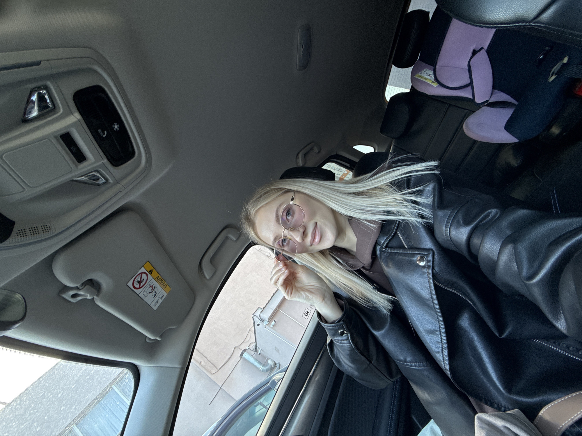 A young, fair-skinned blonde woman with light-framed glasses is seated in the driver's seat of a car. She is wearing a brown hooded sweatshirt under a black leather jacket. The interior of the car is visible, including the sun visor, overhead console with buttons, and a child's car seat in the back. The exterior visible through the car window shows a building with light-colored walls and pipes. Based on the building style, the location is most likely in Izhevsk, Russia.