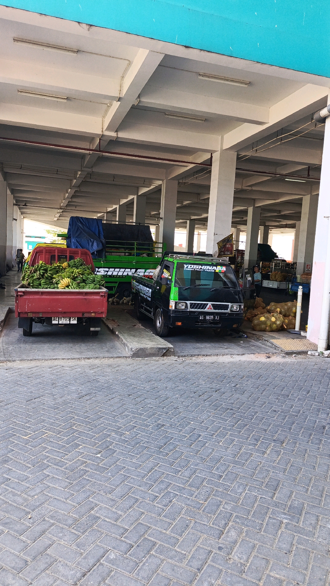 The image depicts an outdoor market scene in Ponorogo, Indonesia. In the foreground, a red pickup truck is loaded with a large quantity of bananas. To its right, a black and green pickup truck with the branding "YOSHINAGA FRUIT TRANS" is parked. Behind these trucks, a larger green truck with a blue tarp is partially visible. The market area is covered by a roof supported by concrete pillars, and the ground is paved with interlocking bricks. Several sacks of produce, possibly bananas, are piled on the ground near the black and green truck. In the background, more market stalls and produce can be seen, along with a few individuals. The overall impression is of a busy fruit market on a sunny day.