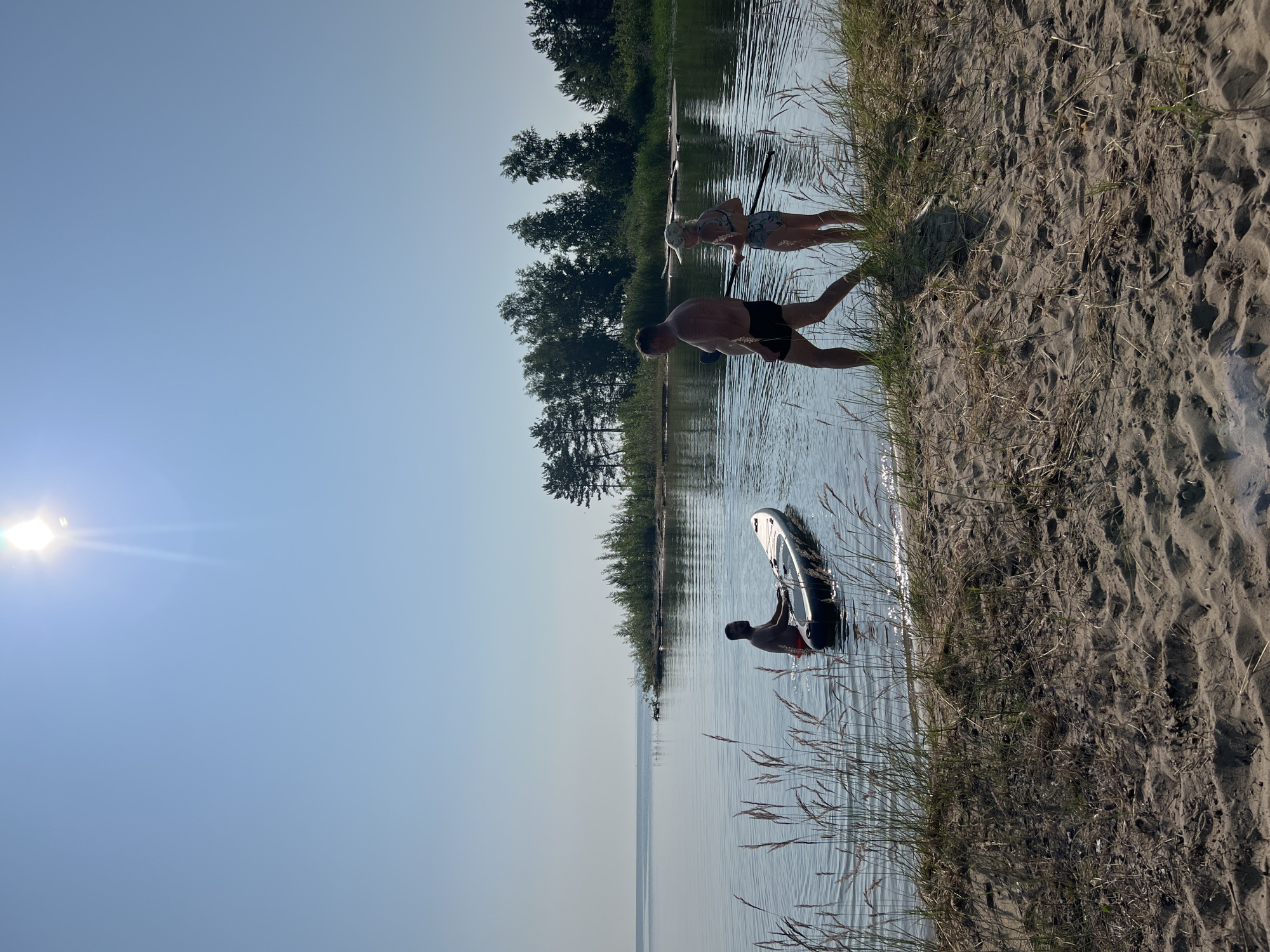 A group of three people are by the water's edge on a sandy beach. The sun is shining brightly in a clear blue sky, suggesting it is daytime. Two individuals, a man and a woman, are partially in the water. The man is facing away from the camera, with his hands in the water, and he is wearing black shorts. The woman is facing the man and is wearing a patterned bikini. Further out in the water, another person, a man, is standing next to a paddleboard, appearing to be getting ready to use it. The background consists of a line of trees. The scene appears to be a recreational activity, likely paddleboarding, taking place on a warm, sunny day in a natural setting.