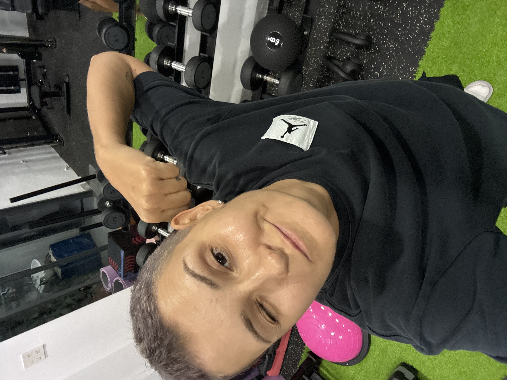 A woman with short, purple-tinged hair is taking a selfie in a gym. She is wearing a black t-shirt with a white Jordan logo on the left chest. She is winking and smiling, with her right arm bent, holding a dumbbell. The gym has a variety of equipment, including weight racks with dumbbells, a black medicine ball labeled "6 KG", and a pink exercise ball on the green floor. In the background, there is a weight machine and a large mirror reflecting more gym equipment and a sink. The scene appears to be located in Nam Nha Trang Ward, Vietnam, indicated by context outside the image.