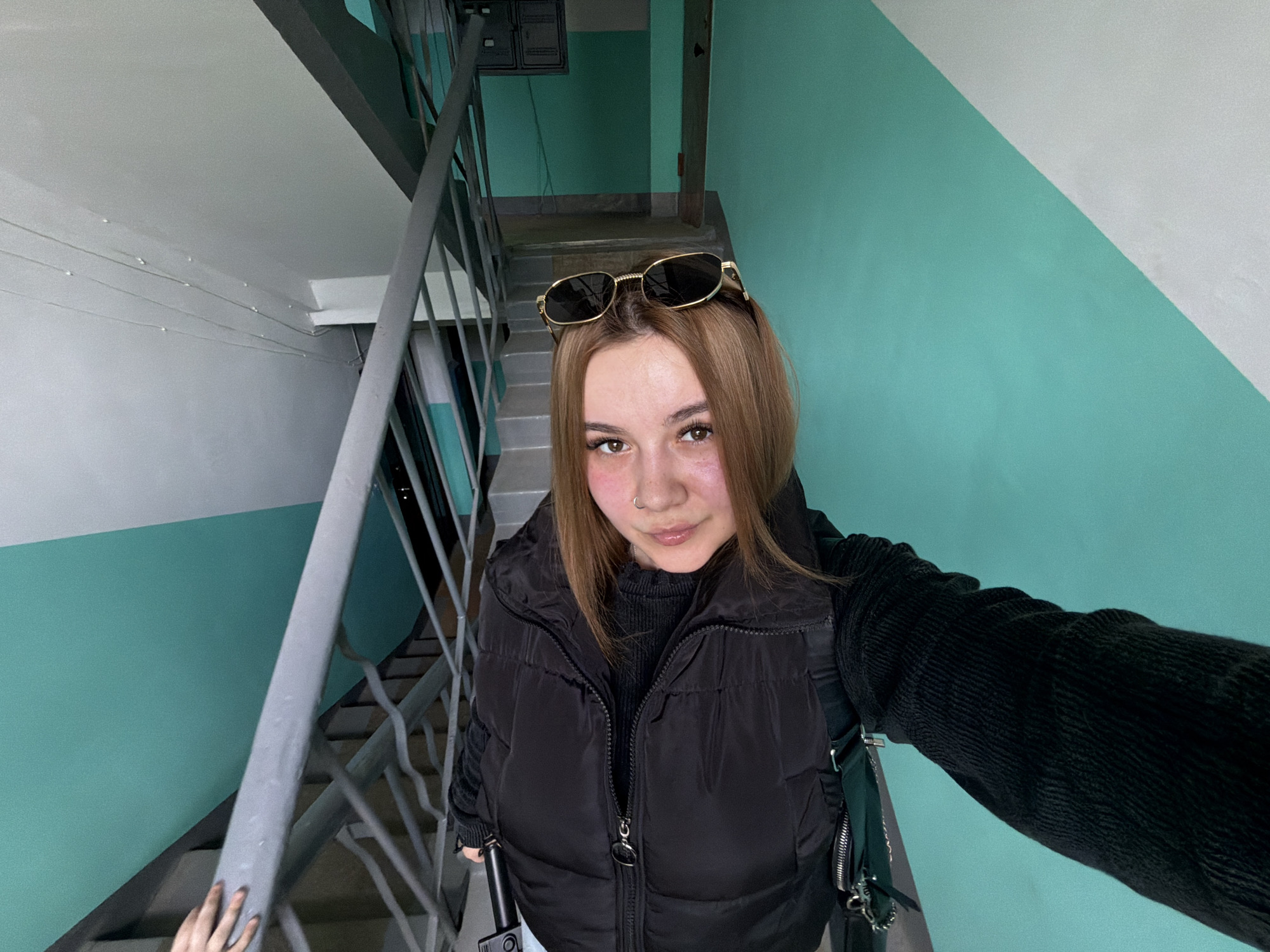 A person is standing on a stairwell, taking a selfie. The stairwell has a teal and white wall, grey railings, and grey stairs. The person has light brown hair and wears a black vest, a black long-sleeved shirt, and sunglasses on their head. They are holding a phone to take the selfie. The location is likely an apartment building stairwell in the city of Chaikovsky, Russia.