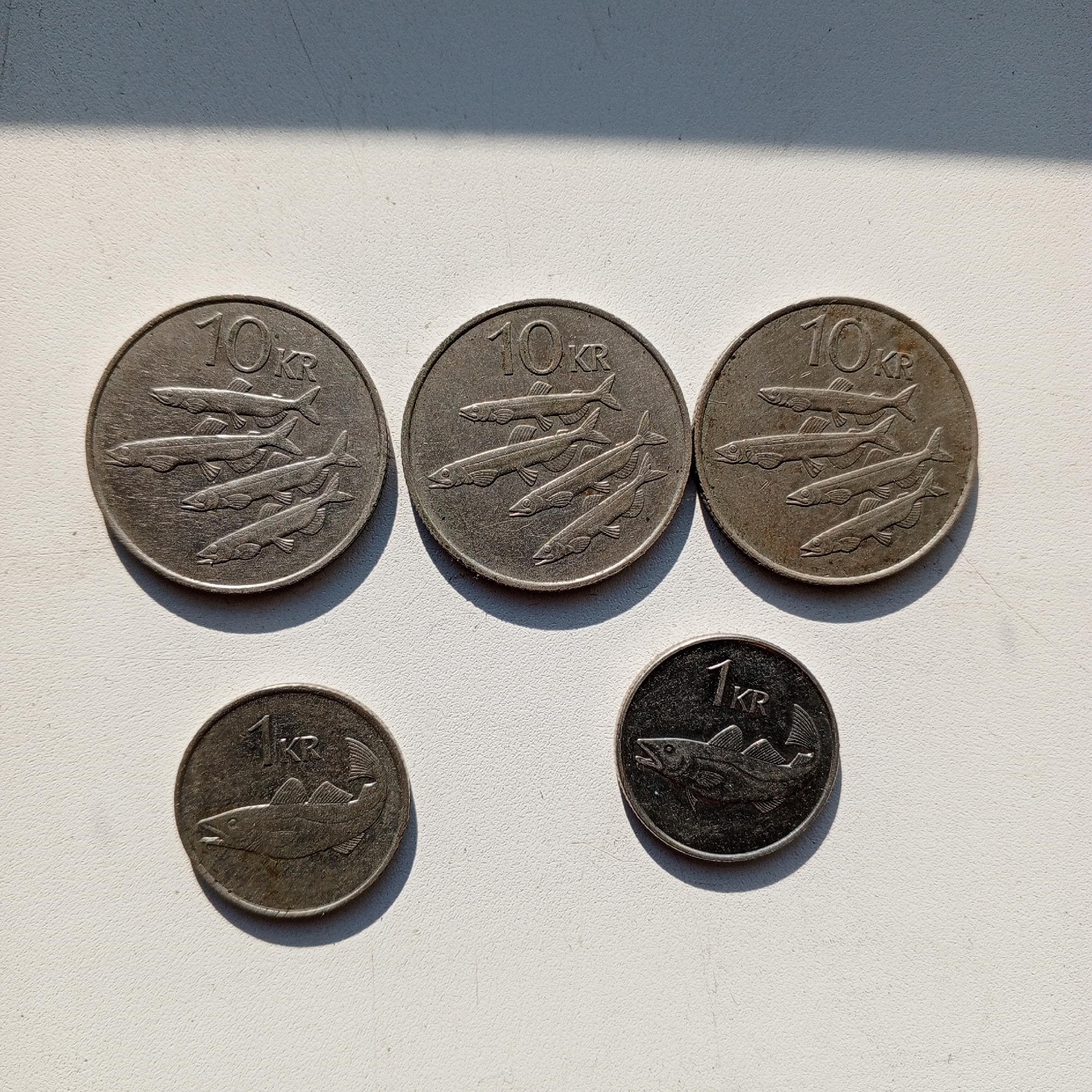 The image displays five coins arranged on a textured white surface, likely a windowsill or table, with a sharp shadow cast diagonally across the upper portion of the frame. Three larger coins, each bearing the inscription "10 KR" and depicting a school of fish, are positioned in the upper half of the image. One coin is on the far left, another in the center, and the third to the right. Two smaller coins, each inscribed with "1 KR" and featuring a single fish, are located in the lower half of the image, with one to the left and another to the right of the center. The lighting suggests it is daytime, possibly morning or late afternoon, given the strong shadow. No people are visible. The coins appear to be old or weathered. The image's focus is on the coins themselves, with the background being a neutral, unidentifiable surface. The context of Sayanogorsk, Russia, is not visually represented in the image.