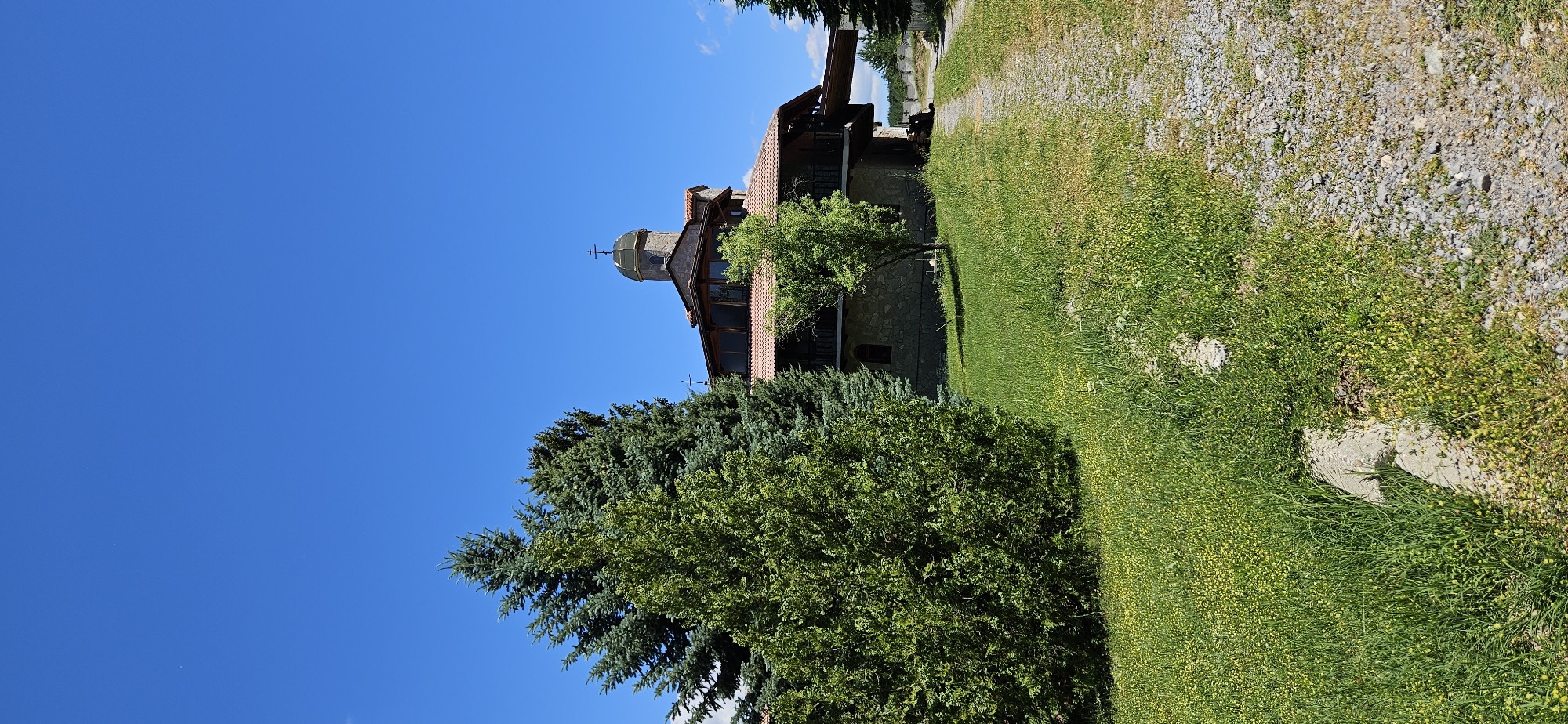 The image depicts a stone building with a red tile roof, topped by a green dome with a cross, suggesting it is a religious structure, possibly a church or monastery. The building is surrounded by lush green grass dotted with small yellow flowers. To the left of the building, there are several large evergreen trees, and a smaller deciduous tree stands in front of the building. A gravel path leads away from the building towards the right side of the frame. The sky is a clear, bright blue. The overall scene is pastoral and appears to be located in a rural area. Based on available information, this location is identified as the Ovchartsi Monastery in the village of Ovchartsi, Bulgaria.