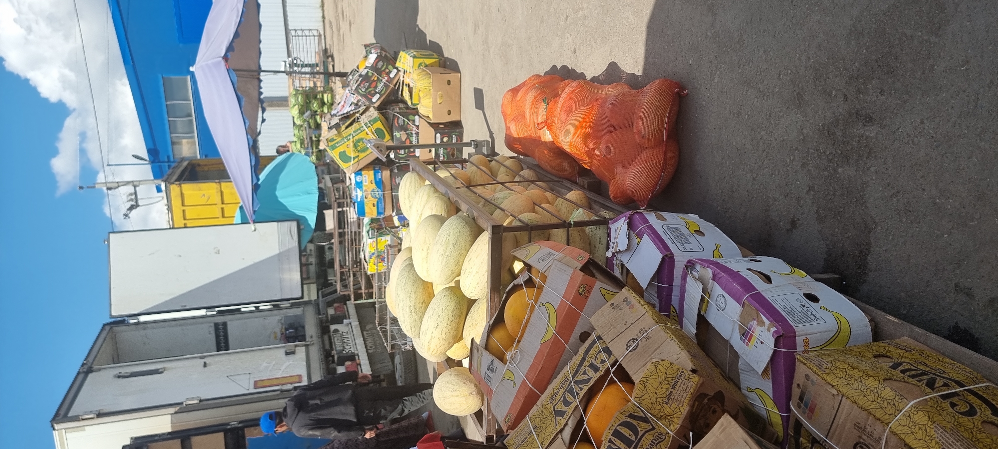 In Aktobe, Kazakhstan, an outdoor market scene unfolds under a bright, cloudy sky. A large white truck with its doors open is parked on the left, a person in a blue cap and black hoodie walks away from it. In the foreground, a metal crate is filled with large, light-colored melons. To the right of the melons, a pile of oranges is contained within an orange mesh bag. Further to the right, stacks of cardboard boxes are visible, some with banana graphics. In the background, a bright blue building stands with a yellow shipping container nearby, and a purple umbrella shades a section of the market. The ground is paved asphalt.