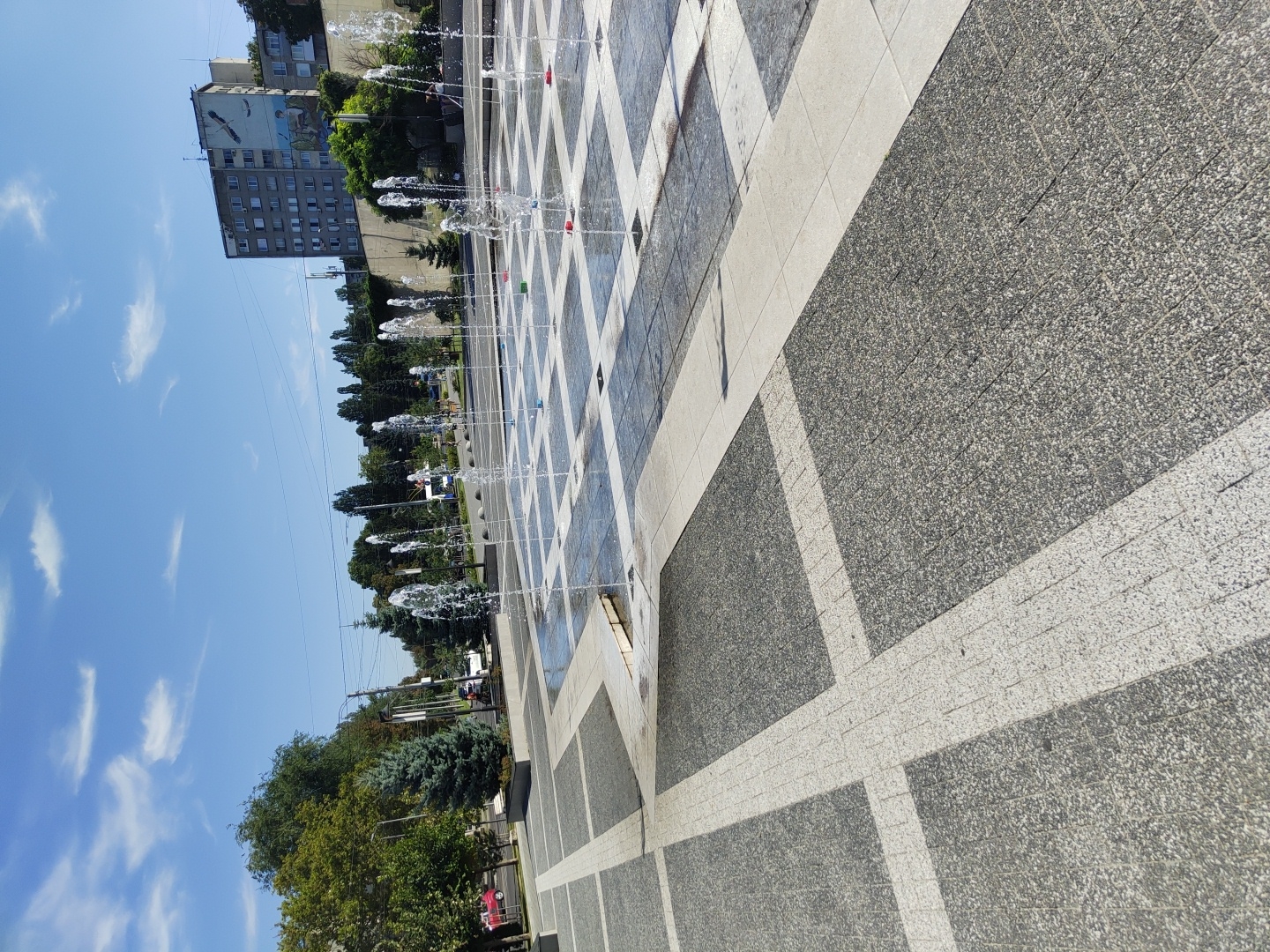 The image shows a public square in Chișinău, Moldova, featuring a fountain with multiple water jets spraying upwards. The ground is paved with a checkerboard pattern of dark grey and white square tiles. The fountain is located in the mid-ground, with water columns rising from individual points on the tiled surface. In the background, there is a multi-story building with a mural on its facade, trees, and a clear blue sky with scattered clouds. The foreground is dominated by the paving of the square. The lighting suggests a sunny day.
