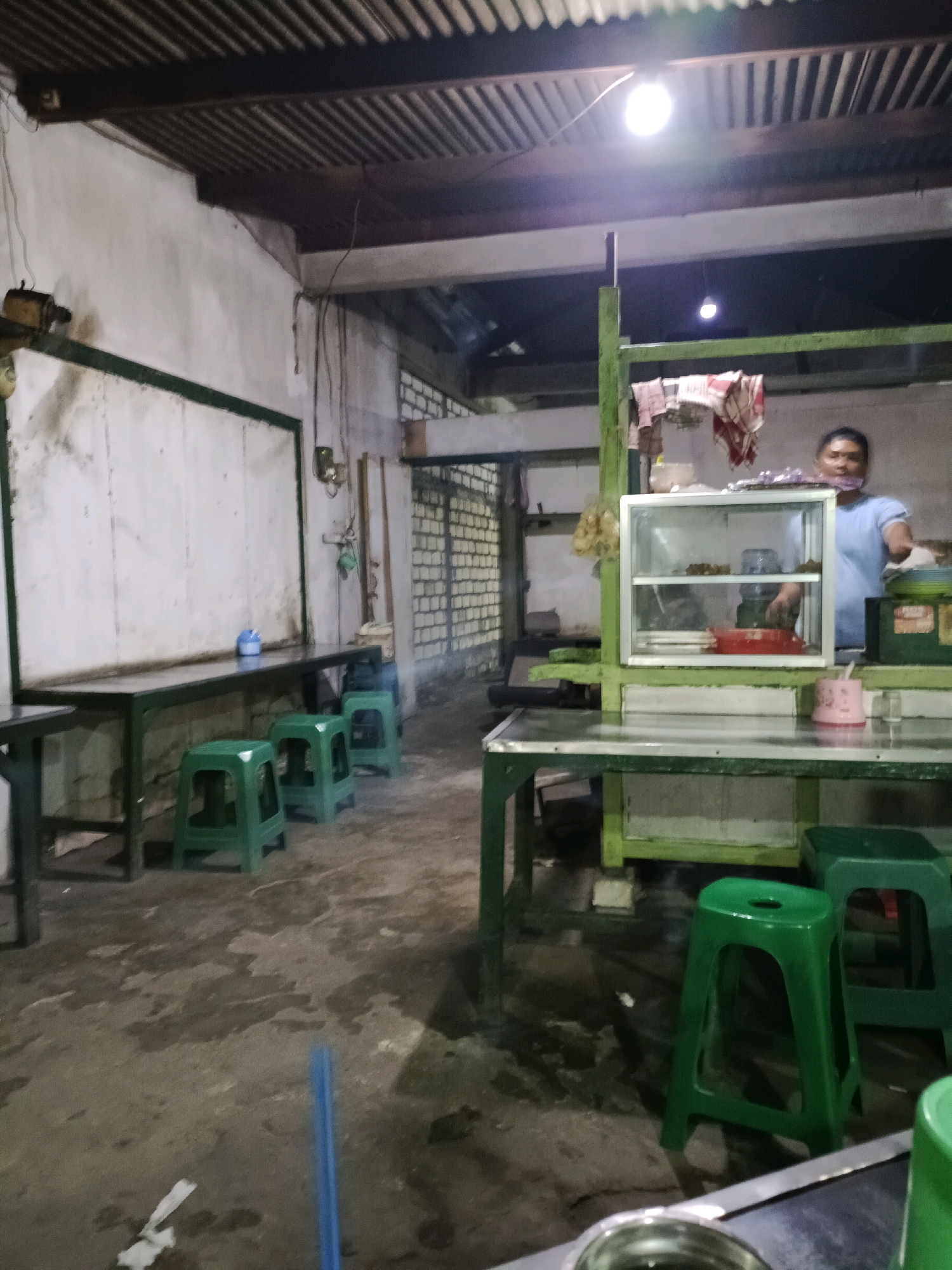 The image depicts an indoor, open-air dining area, likely a street food stall or a simple eatery, in Sukomulyo, Indonesia. The space is lit by a bright, overhead bulb and appears to be a functional rather than decorative establishment. A person, possibly the vendor, is partially visible in the background, wearing a light blue shirt and a face mask. They are behind a counter that features a glass display case filled with various items, and a red bowl is visible on a shelf.
The foreground is dominated by several green plastic stools arranged around simple, dark-colored tables. The floor is concrete and appears wet in places, with some debris scattered on it, including a crumpled piece of paper and a blue plastic straw. In the mid-ground, a long, dark table is set against a white, textured wall, with three green stools neatly placed beneath it. Another table structure, also green, is visible on the right side of the frame, supporting the display case and counter area. The ceiling is corrugated metal, with wooden beams supporting it. The overall impression is of a utilitarian, everyday setting for food service.