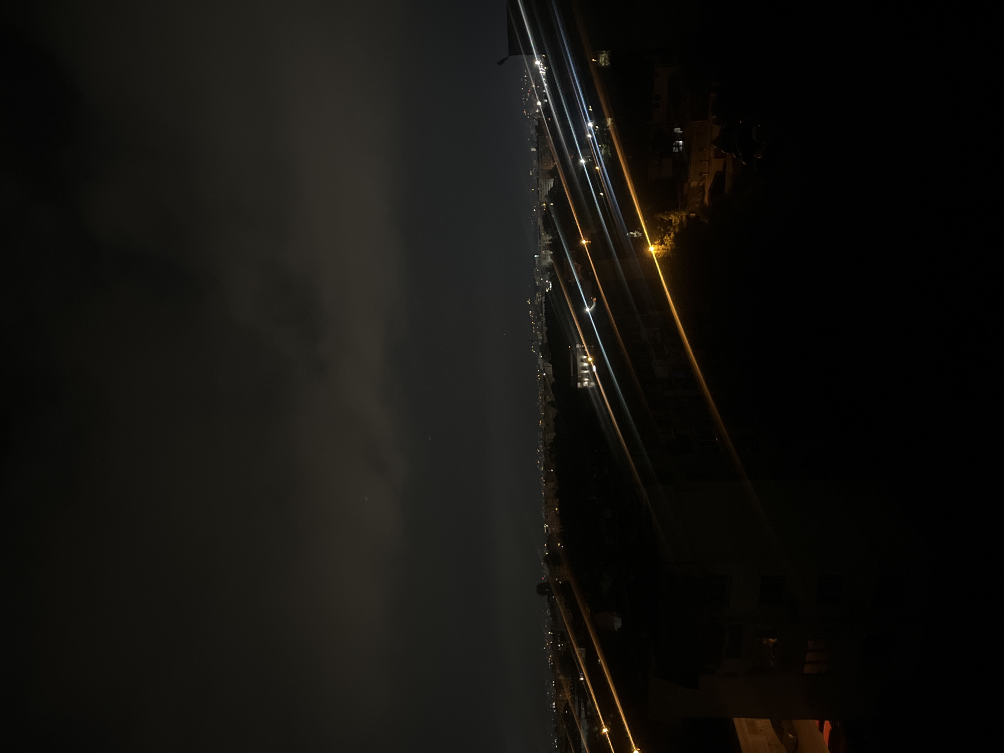 The image shows a nighttime cityscape, possibly in Sofia, Bulgaria, based on the urban environment and building style. The photo is taken from an elevated position, looking out over a city skyline illuminated by streetlights and building lights. There is a heavy fog or cloud cover to the left obscuring the skyline. Linear bright lights likely indicate major streets or highways. There's an architectural structure visible in the foreground, likely a balcony or a building part, adding to the depth of the scene.