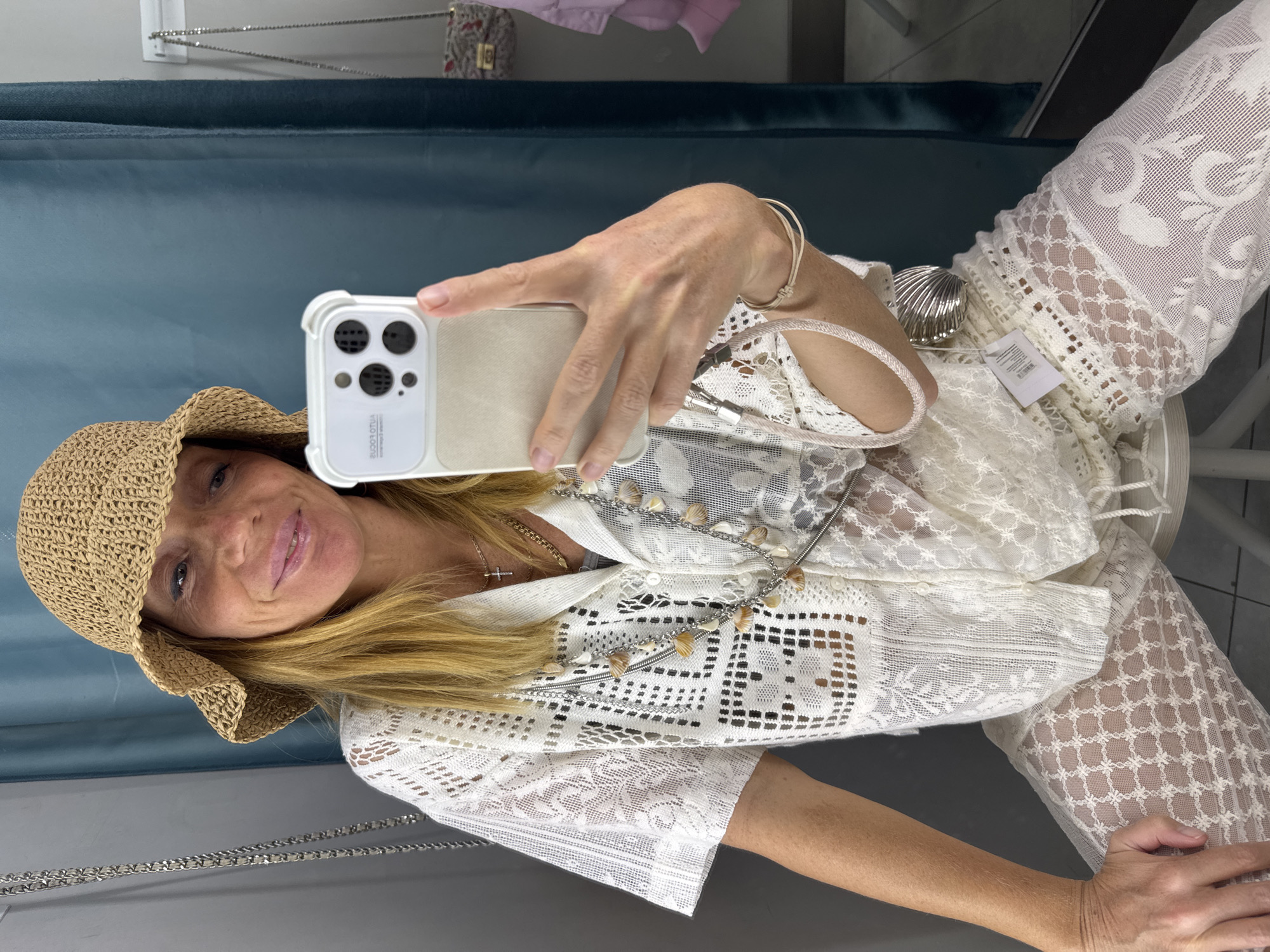 A woman wearing a straw hat, white lace top, and pants is taking a selfie with her smartphone in a room with teal-colored walls. She has long blonde hair and is smiling. A silver purse chain is visible along the edge of the wall. A silver seashell ornament is partially visible in the background. It is not possible to determine the location of this scene in Saint Petersburg, Russia based on the image.