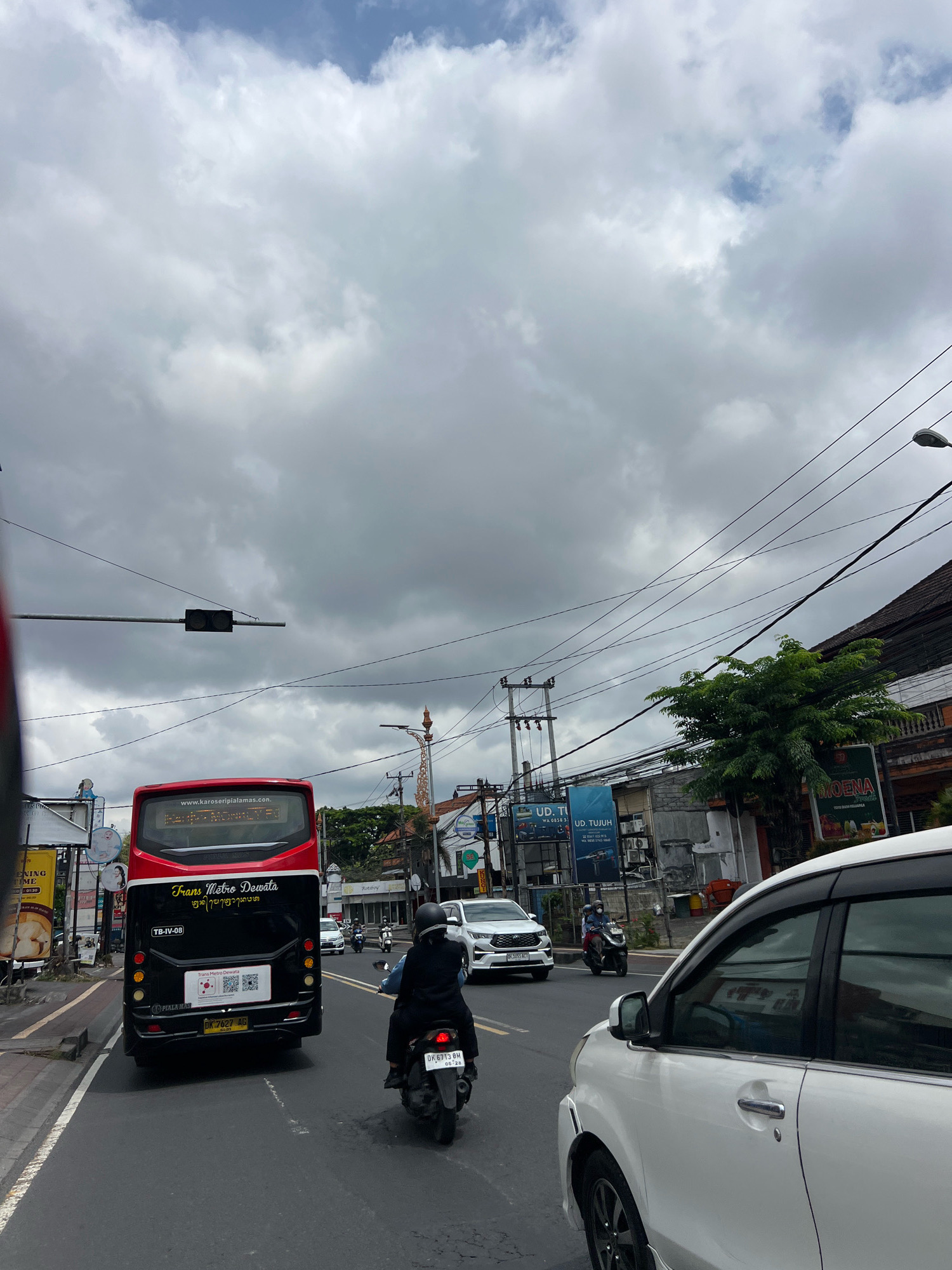This is a street-level, eye-level, medium shot of traffic on a cloudy day in Denpasar, Indonesia. The perspective is from inside a moving vehicle, looking forward. The immediate foreground is occupied by the right side of a white car. Beyond the white car, in the middle ground, is a red and black bus labeled "Trans Metro Dewata" with license plate DK 7627 AG. To the right of the bus is a person riding a motorcycle, wearing black clothing and a black helmet. Further in the background, traffic continues with more cars and motorcycles. Buildings with signs and trees line the right side of the street. Overhead, power lines crisscross the sky, which is filled with heavy, grey clouds, indicating an overcast or impending rainy day. The street appears to be paved and marked with white lines. The overall impression is a typical urban scene in Denpasar, characterized by active traffic and a humid, tropical atmosphere.