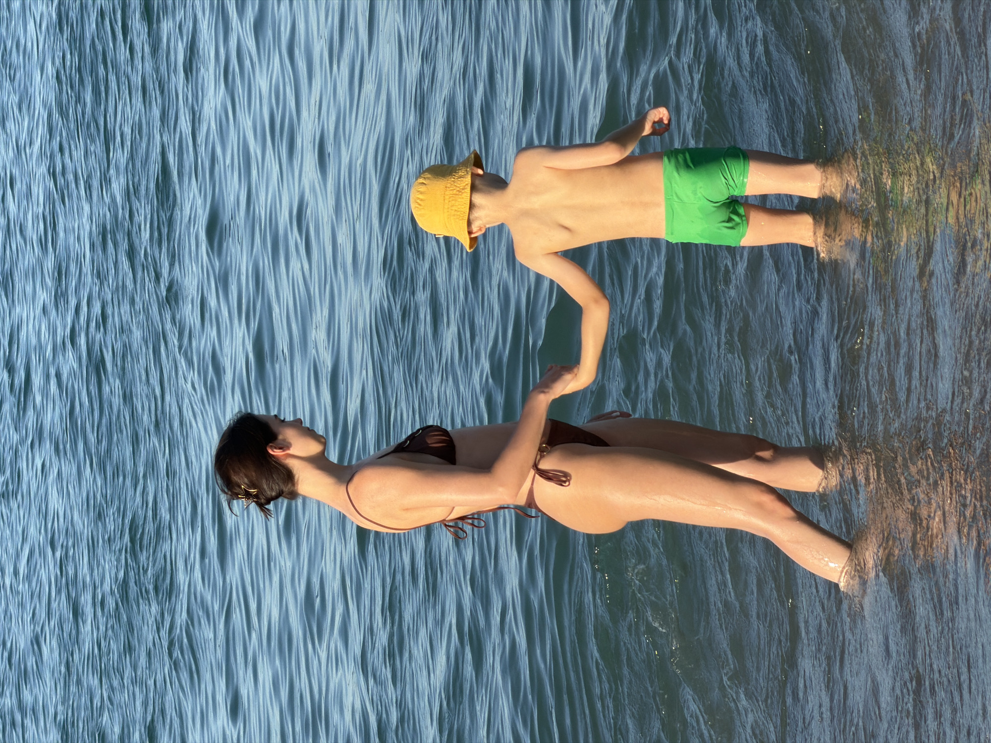 Two people, an adult and a child, are standing in shallow, rippling blue water. The adult, a woman wearing a brown bikini, is in the foreground, facing away from the camera. Her head is tilted back, and her arms are slightly raised, with her hands near the child's. The child, wearing a yellow bucket hat and green swim trunks, is in the background, facing the woman. The child's left arm is extended towards the woman, and their right arm is raised. Sunlight glints off the water, suggesting it is daytime, likely sunny given the bright reflections. The scene appears to be a beach or a shallow body of water, possibly the Mediterranean Sea given the Barcelona context. No text is visible in the image. The overall impression is one of leisure and a shared moment between the two individuals.