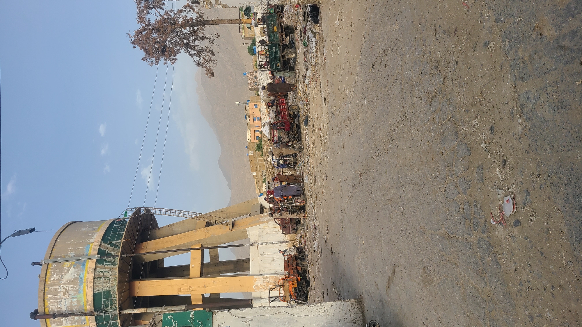 The image captures a scene in Quetta City Tehsil, Pakistan. On the left, a cylindrical water tower stands prominently, supported by concrete pillars. A network of ladders and walkways encircles the tower's exterior. Overhead, power lines stretch across the sky. A row of auto rickshaws lines the edge of the road, with people gathered around them. In the background, a mountain range forms the backdrop, and buildings are scattered. The road is asphalt, showing signs of wear and tear.