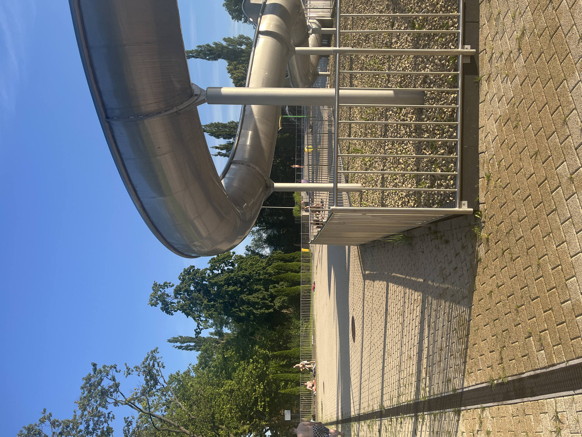 A wide shot captures a curved, metallic water slide at a public park or water park under a clear blue sky. The slide dominates the right side of the frame, curving from the top right downwards and to the left. A metal fence encloses the base of the slide, where small rocks and sparse green vegetation are visible. The foreground is paved with light-colored rectangular bricks, with a dark metal grate running along the left edge.
In the background, beyond the fence and some trees, several people are scattered across a paved area. Some are sitting or lying on towels, while others appear to be standing or interacting. These individuals are mostly in swimwear. The environment is lush with green trees, some tall and slender, others full and bushy, forming a natural backdrop. The scene is bathed in sunlight, casting long shadows from the slide and fence onto the paved ground. The overall impression is a sunny, recreational outdoor setting.