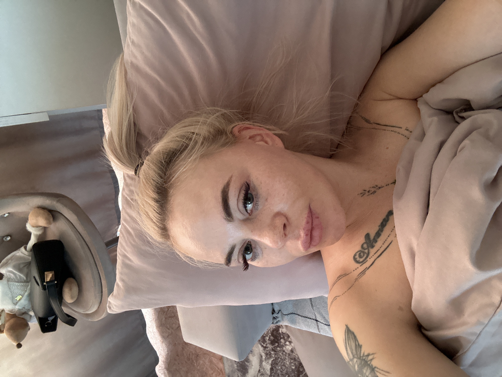 A woman with blonde hair pulled back in a ponytail lies on a pink pillow covered by a pink blanket. She has tattoos on her chest and shoulder. A small chair with a stuffed animal sits to her left. The location information is not determinable from the image.