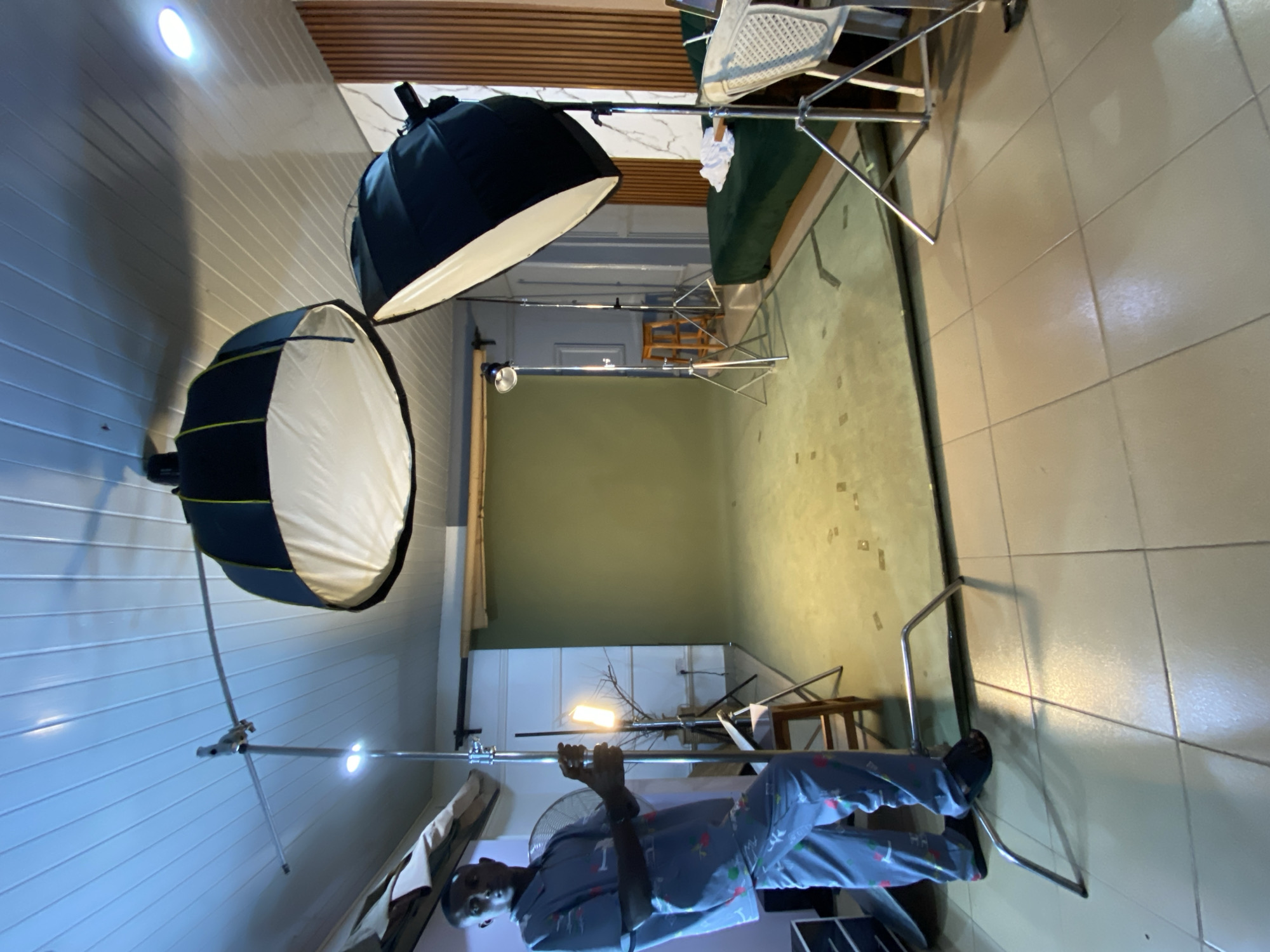 This image captures an indoor scene that appears to be a makeshift photography studio, possibly located in Bukuru, Nigeria. The room features a light green wall or backdrop on the left side, extending downwards to a light-colored tiled floor. The walls to the left and the ceiling are white and have horizontal paneling.
In the foreground, a man stands centrally, facing the camera. He is dressed in light blue or grey pajamas with small, colorful, scattered patterns. He holds onto a horizontal metal bar, which is part of a light stand setup. A small fan is visible behind him.
The space is equipped with several pieces of photography lighting equipment. Two large softboxes are prominent on the left, mounted on tall metal stands. One is a round parabolic softbox, and the other is a rectangular softbox, both featuring white diffusers and black exteriors. Another bright, bare light is visible on a stand further back, pointed towards the green backdrop. Numerous metal light stands and support bars are distributed across the room, suggesting a comprehensive lighting arrangement.
In the background, on the right side of the green backdrop, a dark green sofa or bench is visible. Above it, an inverted white chair with a woven pattern rests on what appears to be another prop or stand. Additional wooden furniture, possibly a stool or table, can be seen further into the background on the right. The active artificial lighting indicates a controlled environment for a photoshoot. No visible text is discernible in the image.