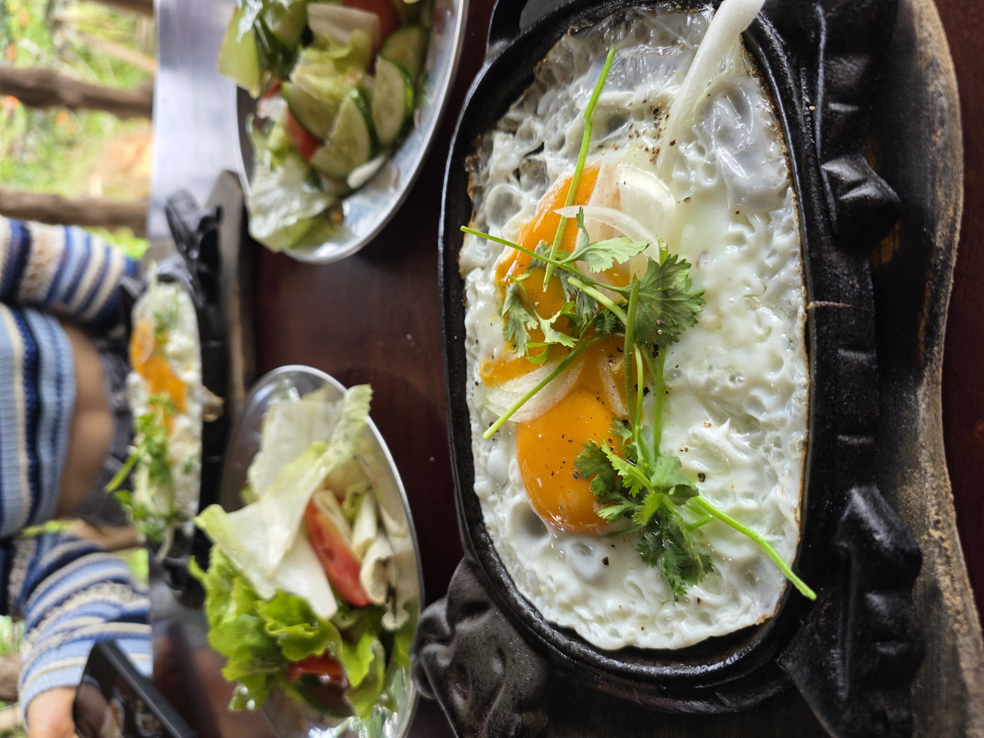 This media file captures a close-up, high-angle view of a meal being served outdoors during the daytime in Sơn Điền, Vietnam.
The central focus is a black cast-iron sizzling plate holding a fried egg. The egg features a perfectly cooked white with slightly crispy edges and a vibrant, sunny orange yolk, possibly indicating two yolks. It is garnished with fresh green cilantro/coriander sprigs, thin slices of white onion, and a sprinkle of black pepper.
Accompanying the main dish are two clear glass bowls filled with fresh green salad. Each bowl contains crisp green lettuce, slices of cucumber, and chopped red tomatoes. One of the salads also appears to have some light-colored, possibly sliced mushrooms or another vegetable.
A person is partially visible on the left side of the frame, wearing a blue and light-colored striped long-sleeved sweater. Their hand is holding a black smartphone, suggesting they might be taking a photo of the meal or engaging with their device during the dining experience.
The meal is laid out on a dark brown table. The background, largely out of focus, shows lush green foliage and what appears to be wooden railings or a fence, indicating an outdoor or semi-outdoor dining setting, possibly on a patio or balcony. The natural lighting suggests it is daytime. No text is visible in the image. The arrangement of the food suggests a casual and inviting meal, likely breakfast or brunch.