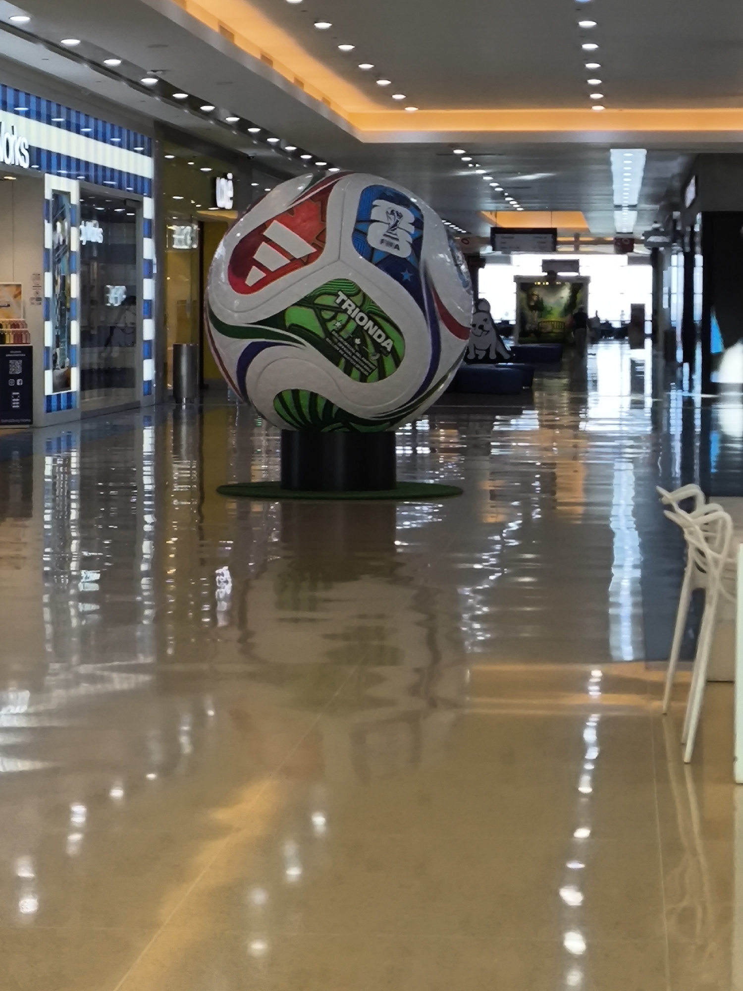 The image shows a large soccer ball sculpture in a shopping mall in Porlamar, Venezuela. The ball is painted with the Adidas logo, the FIFA logo, and the word "Trionda." It sits on a black base with a green ring at the bottom. The mall's floor is shiny and reflects the lights and the sculpture. In the background, there is a store on the left and some seating area on the right. Some chairs are visible in the lower right corner of the image. The scene is brightly lit by overhead lights.