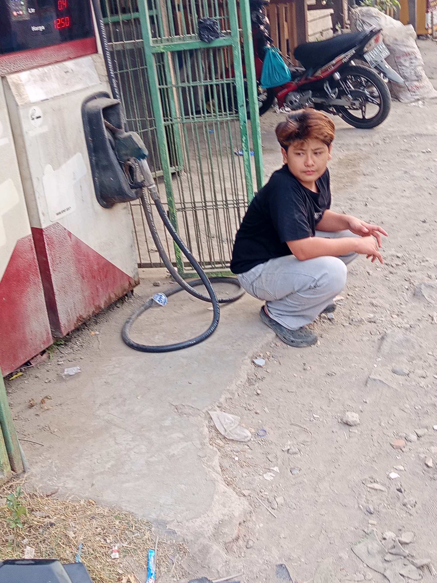 A young person with short, light brown hair is squatting on a dusty, concrete ground in what appears to be an outdoor setting. They are wearing a black t-shirt and light gray pants, with dark clogs on their feet. Their hands are positioned in front of them, palms down, with fingers slightly curled.
In the background, to the left, is a red and white gas pump with a black nozzle and hose. The nozzle is resting in its holder. The gas pump has a digital display showing "0.4" and "12.50". A green metal gate is visible behind the gas pump, and beyond that, a motorcycle is parked. A blue bag is hanging from the back of the motorcycle.
The ground is a mix of dirt, gravel, and scattered debris, including small pieces of plastic. The overall environment suggests an outdoor location, possibly a small gas station or a similar service area in Karawang, Indonesia. The lighting indicates it is daytime.