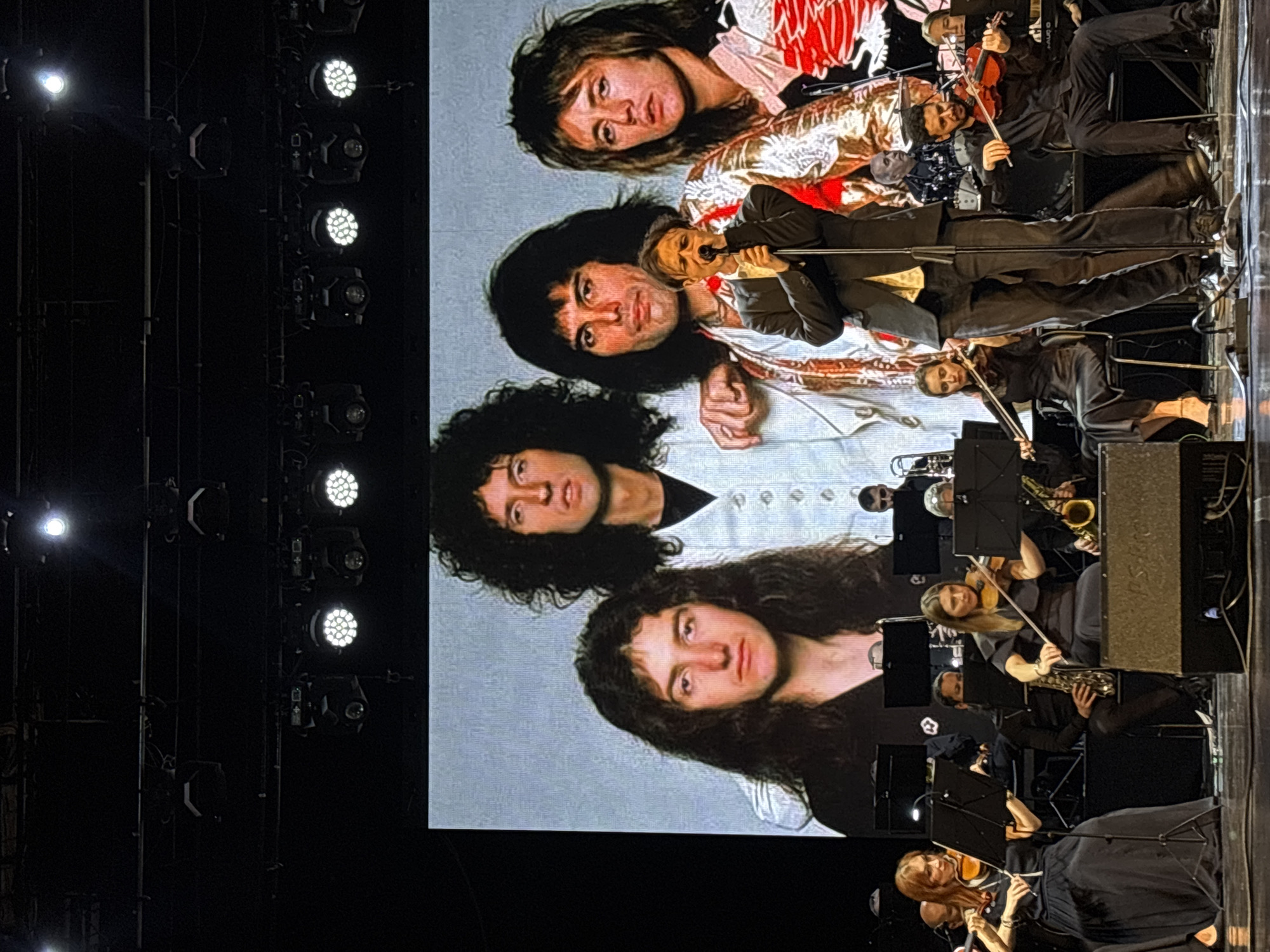 A large screen displays a group photo of the rock band Queen, consisting of Freddie Mercury, Brian May, Roger Taylor, and John Deacon. The screen is positioned to the left of an orchestra performing on stage. The orchestra members are dressed in black. The venue appears to be an indoor concert hall. The performance location is Chaikovsky, Russia.