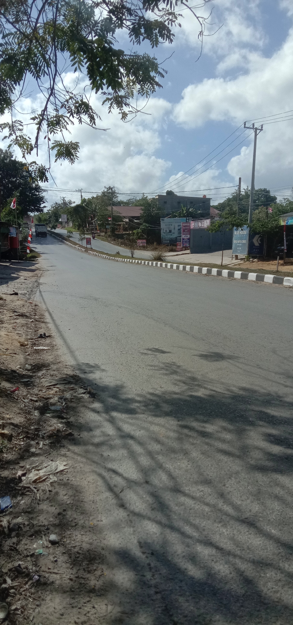 The image captures a street scene in Kendari, Indonesia, under a partly cloudy sky. A paved road stretches into the distance, curving slightly to the left. The road is bordered by a white and black striped curb on the right. On the left side of the road, there is a dirt shoulder with scattered debris. Lush green trees with dense foliage line the left side of the road, some branches extending over the pavement. On the right side of the road, there are buildings and various signs, including one that reads "BLUE HOUSE" with a phone number. A utility pole with power lines is visible on the right. Shadows of tree branches are cast across the road. In the distance, a truck is seen driving on the road, and a motorcycle is parked on the side. The overall impression is of a moderately busy street in a tropical urban environment.