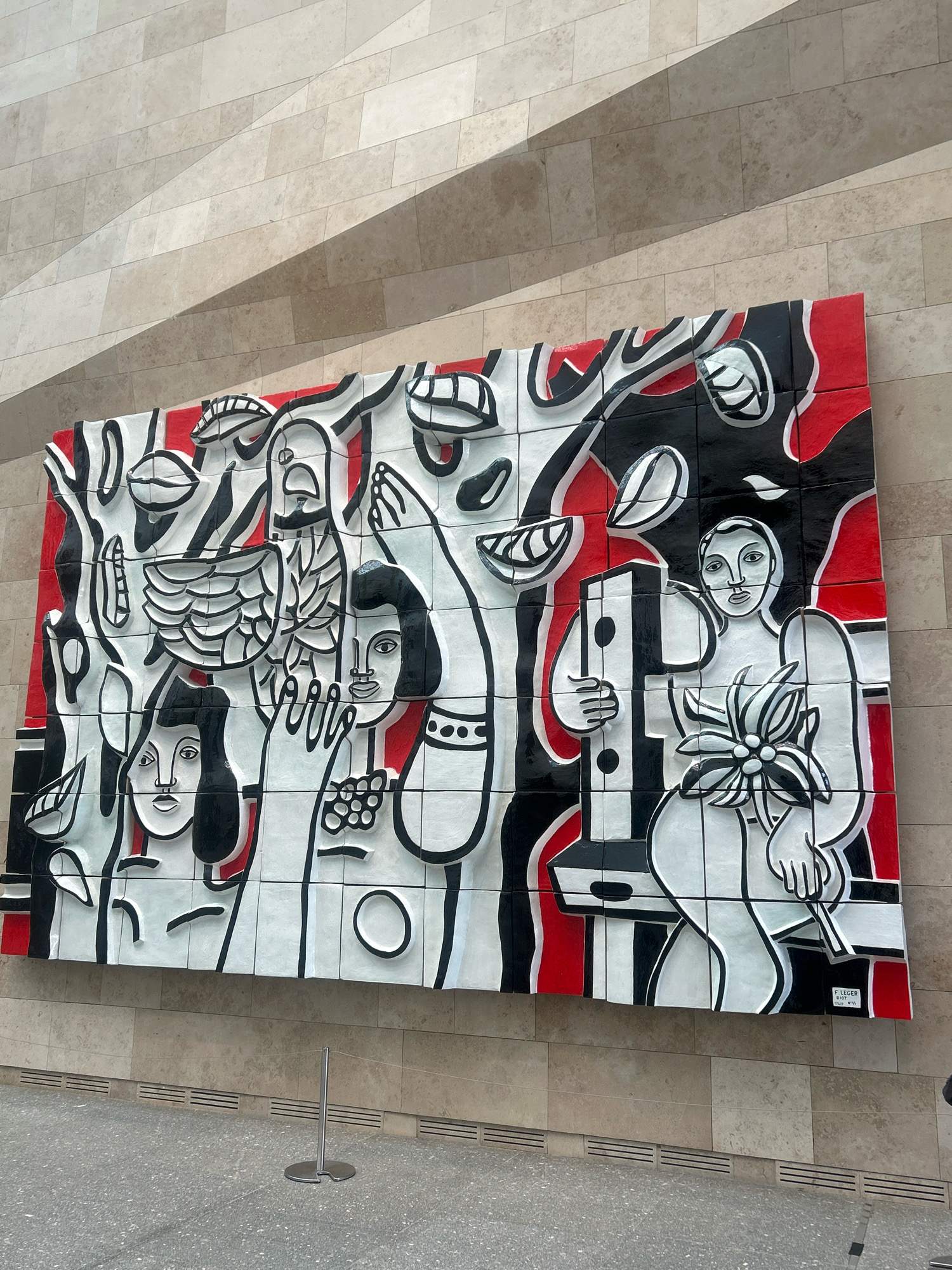 The image shows a large, colorful mural mounted on a light-colored, tiled wall. The mural is abstract and features stylized figures and organic shapes rendered in bold black outlines against a background of white and red. The artwork appears to be made of ceramic tiles. In the foreground, the floor is a speckled gray material, and a thin metal stanchion with a wire is visible. The wall behind the mural is constructed of rectangular stone tiles in varying shades of beige and light brown, with a diagonal shadow cast across it. The lighting suggests an indoor setting, possibly a public building or gallery. The mural is signed in the lower right corner with "F. LEGER BIOT 1952-1954", indicating it is likely a piece by Fernand Léger and Marcel Biot. This artwork is located at the Central State Museum of Kazakhstan in Almaty, Kazakhstan.