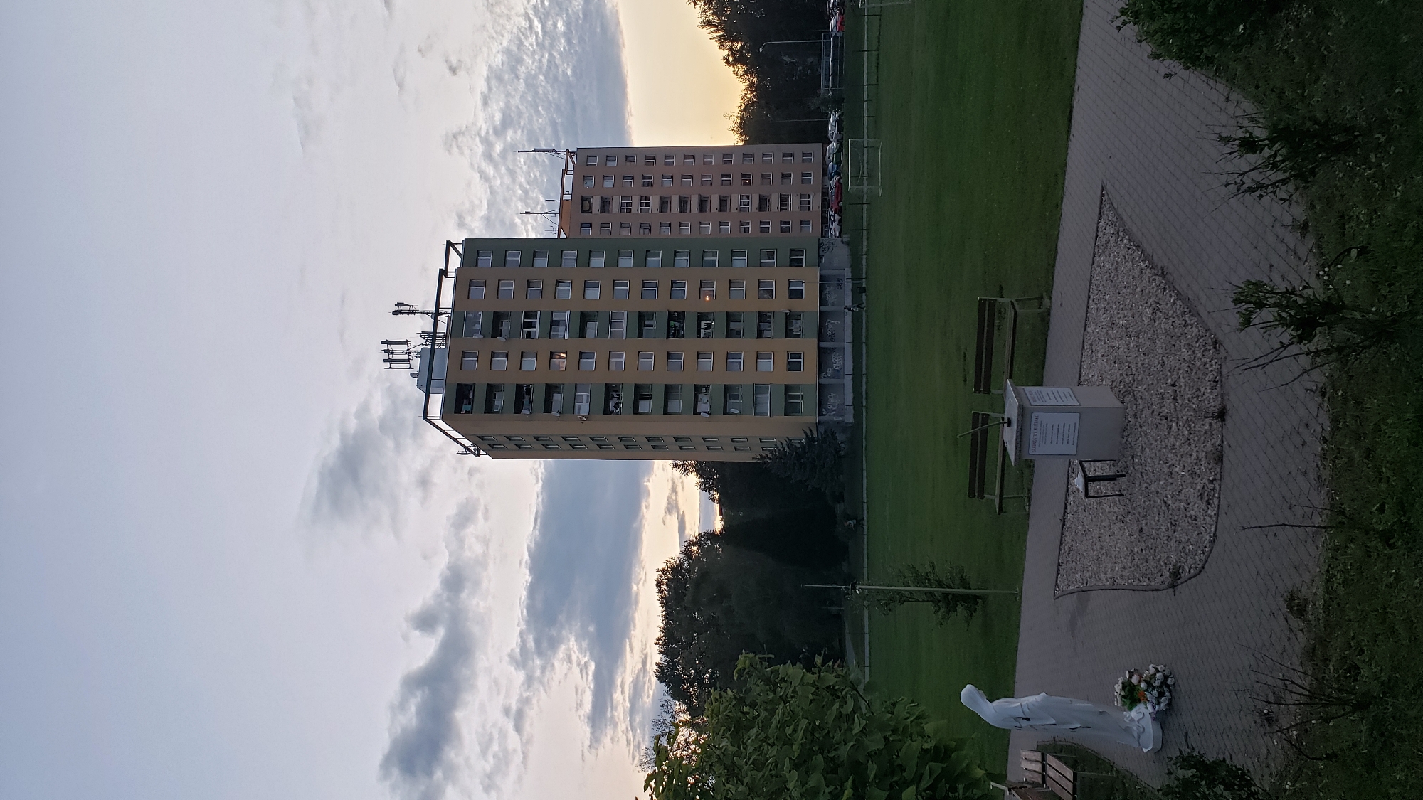 In Prešov, Slovakia, an outdoor scene captures two identical residential buildings situated side-by-side, with one slightly recessed behind the other. The buildings are tall, multi-story structures with a light yellow facade and green accents. Both are topped with communication antennas. In the foreground, a paved pathway leads to a small, gravel-filled area with a statue of a person in white, adorned with flowers. Adjacent to this, a small monument stands with a plaque. Further back, a manicured lawn stretches out, dotted with scattered trees and two park benches. The sky above is a gradient of twilight hues, with scattered clouds and the warm glow of sunset visible. A parking lot with several cars is visible behind the buildings, and a football field with goalposts is also present on the lawn. The overall atmosphere is tranquil and subdued.