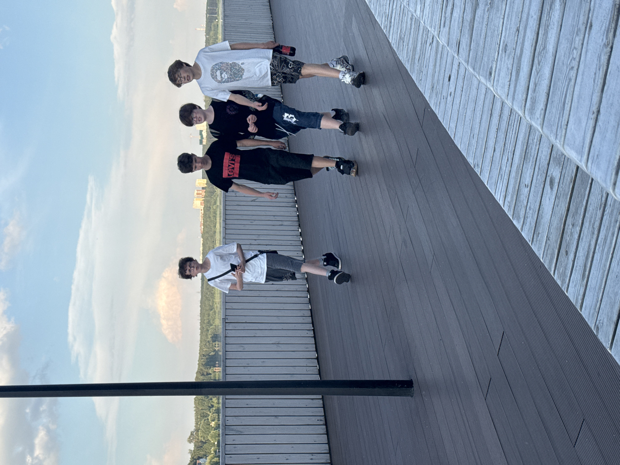 Four young males are walking in single file on a dark wooden boardwalk next to a pale wooden railing. The male on the far left is wearing glasses, a white t-shirt, and grey shorts. He is holding a black smartphone in his left hand and has a black crossbody bag over his right shoulder. The second male from the left is wearing a black t-shirt with red lettering and black shorts. The third male from the left is wearing a black t-shirt and denim shorts. The male on the far right is wearing a white t-shirt with a graphic design and patterned shorts. He is holding a red bottle in his right hand. In the background, there is a body of water, trees, and buildings, suggesting an urban waterfront setting. The sky is partly cloudy with a pale blue hue. This scene is likely located on the embankment of the Iset River in Yekaterinburg, Russia, specifically along the Plotinka embankment, which features wooden boardwalks and offers views of the city.