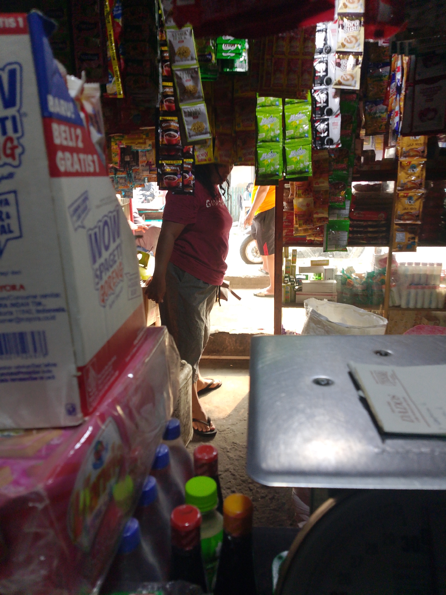 The image depicts a small, cluttered shop, likely a "warung" or convenience store, located in Palembang, Indonesia. The foreground is dominated by a portion of a metal counter, on which a piece of white paper with text is visible. To the left of the counter, a large cardboard box, possibly for selling food items like spaghetti, is prominently displayed. The box has blue and white branding, and text that includes "WOW SPAGETTI". Behind the box, several rows of bottles with colorful caps (blue, red, green, yellow) are stacked.
In the mid-ground, a woman wearing a maroon t-shirt and grey trousers stands facing towards the right, her left hand resting near a shopping bag. Another person, wearing a bright yellow t-shirt and dark shorts, is partially visible in the background, standing near a motorcycle.
The shop's interior is densely packed with goods. Shelves are laden with various packaged food items, snacks, and drinks, many of which are hanging from the ceiling. Products include packets of coffee, tea, biscuits, and other processed foods. The lighting suggests it is daytime, with sunlight filtering in from the open entrance, illuminating the scene. The overall impression is of a typical, small-scale retail environment in a South Sumatran urban setting.
