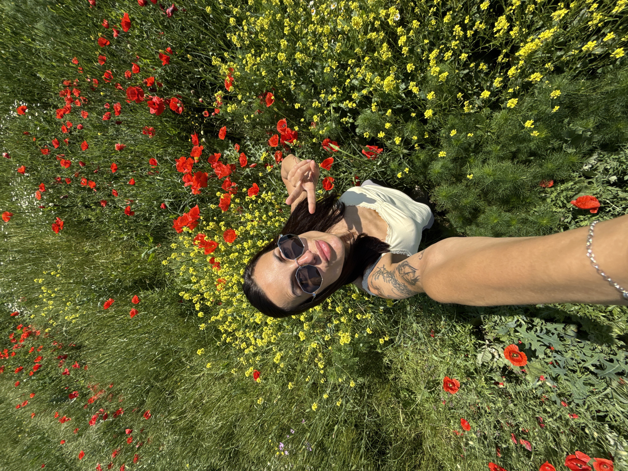 A high-angle view shows a person lying in a field of wildflowers, taking a selfie. The field is filled with red poppies, yellow wildflowers, and green foliage. The person has dark hair and is wearing sunglasses and a light-colored top. A tattooed arm extends into the frame, holding the camera. The location is a field in Pleven, Bulgaria.