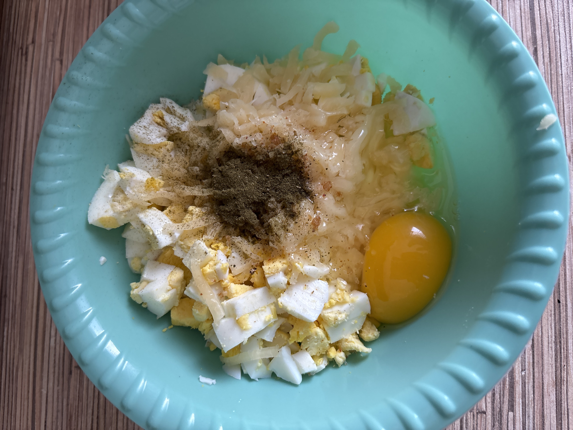 A close-up, overhead shot shows a light teal bowl containing chopped hard-boiled eggs, shredded cheese, a raw egg yolk, and a pile of ground black pepper. The ingredients are mixed together, suggesting preparation for a dish. The bowl is placed on a wooden surface with a visible grain pattern. There is no indication of the location being Naberezhnye Chelny, Russia, as this is a generic kitchen setting.