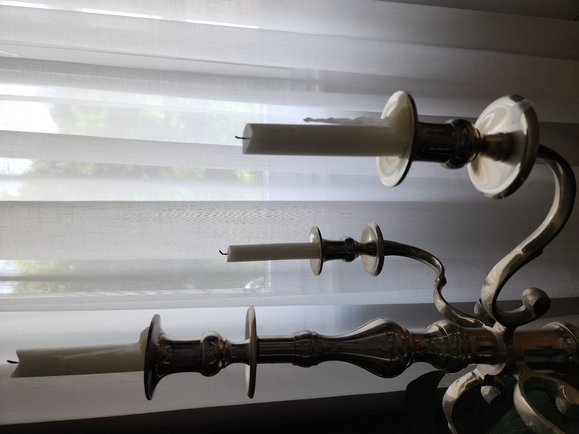 The image depicts an unlit, three-branched metallic candelabra, likely brass or a gold-toned alloy, positioned indoors in the city of Idar-Oberstein, Germany. Three white, cylindrical pillar candles are seated in the candelabra's holders. The candle on the far left is the tallest, appearing largely unburnt. The middle candle is significantly shorter, while the candle on the right is of medium height and exhibits visible wax drips along its side, indicating prior burning. All three candles feature exposed black wicks and are currently unlit. The candelabra itself is highly reflective, showing highlights and shadows across its ornate, polished surface, which features a central stem and three curved arms. In the background, a large window is obscured by multiple vertical panels of white, sheer, translucent curtains. Diffused natural light permeates through these curtains, revealing indistinct green foliage, likely trees or bushes, outside the window, suggesting a daytime scene. The overall lighting creates a backlight effect, highlighting the translucency of the curtains and casting the candelabra in partial silhouette with strong reflections. No individuals are present in the scene.