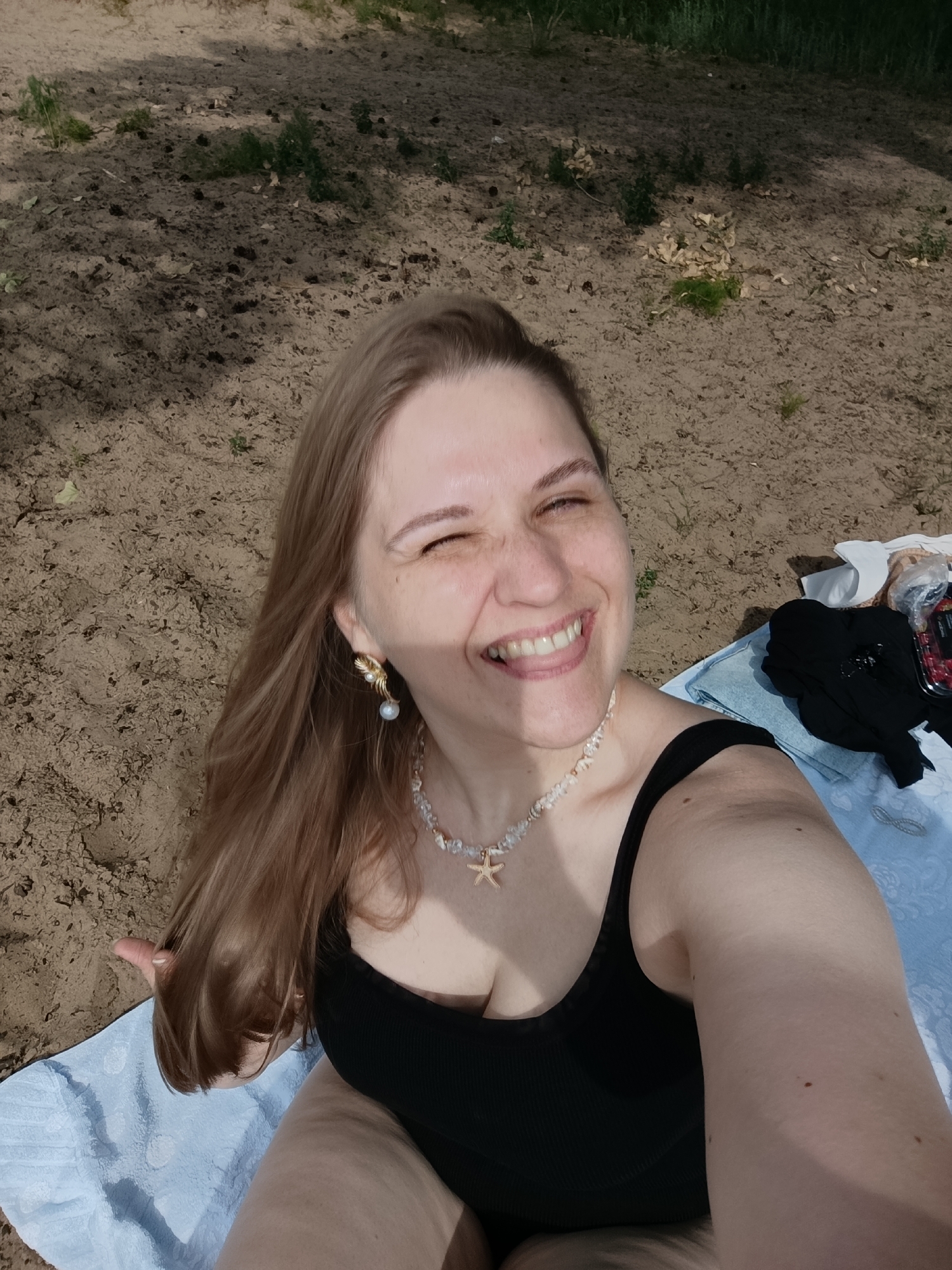 A light-skinned woman with long, blonde hair and fair skin is taking a selfie on a sandy beach. She is wearing a black swimsuit and a seashell necklace, and a pearl earring adorns her left ear. Her right eye is closed in a wink, and she is smiling broadly, revealing her teeth. A light blue towel is spread out beneath her, and some clothing and a container of berries are visible to her right. The background consists of sand, scattered leaves, and shadows cast by trees. The overall scene suggests a sunny day outdoors.