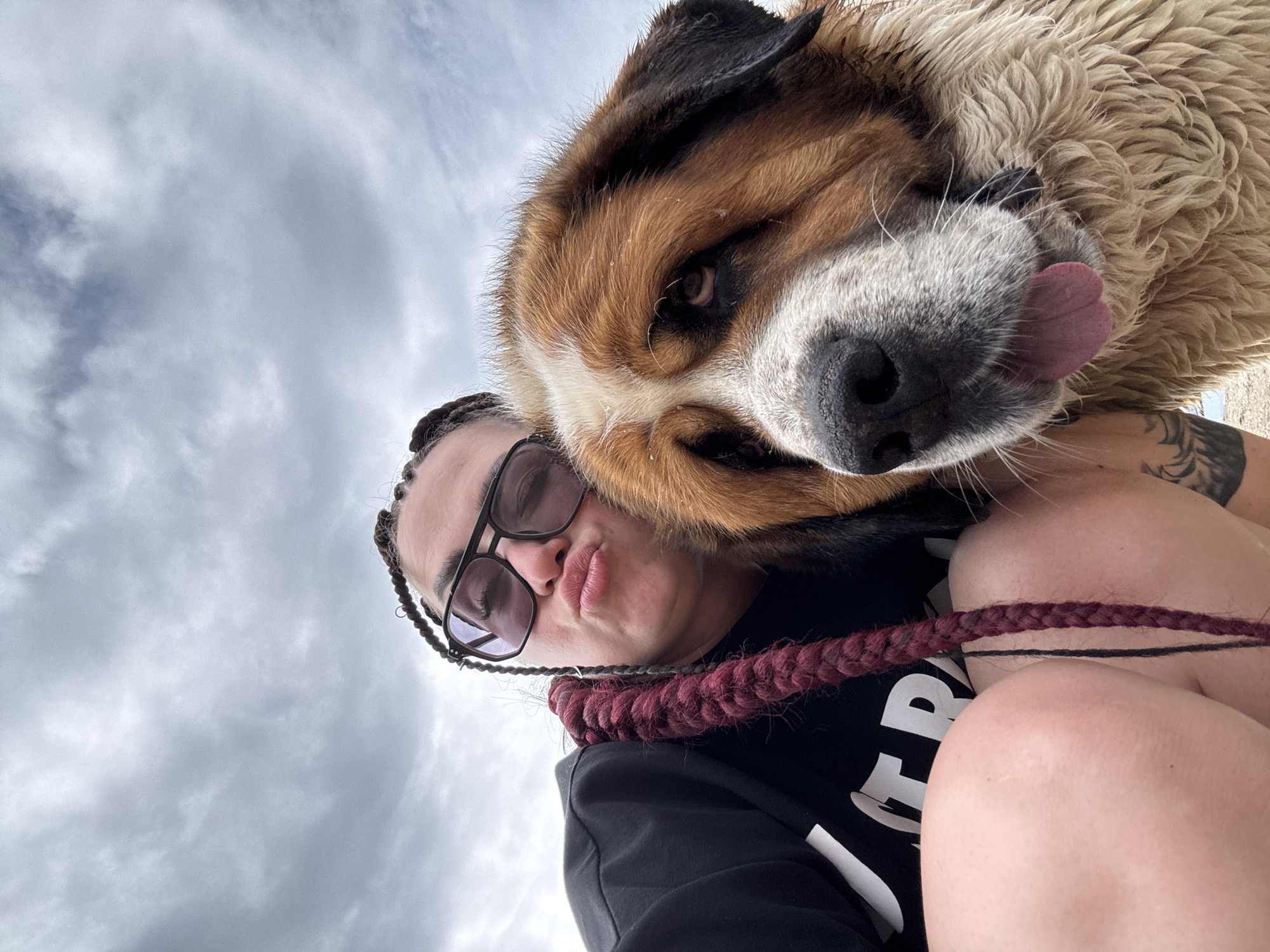 A low-angle, close-up selfie shows a person with dark skin and long, burgundy braids wearing sunglasses and a black sweatshirt. The person is puckering their lips and holding a large Saint Bernard dog, whose head is also in the frame. The dog's fur is wet, and its tongue is sticking out. The background is a cloudy sky.