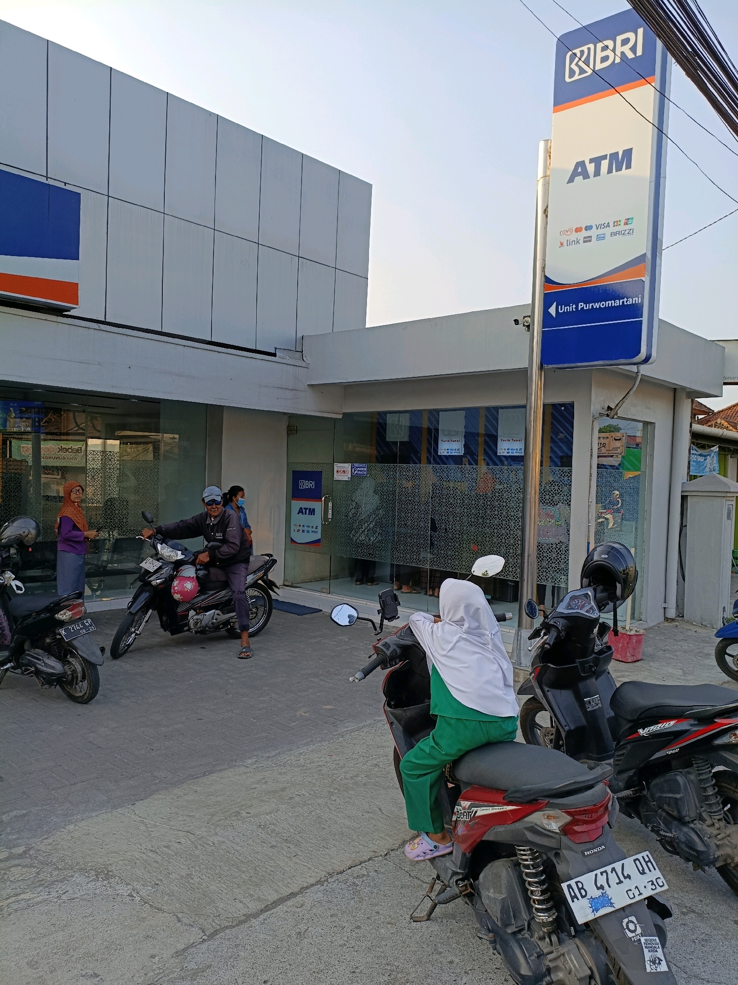 The image shows the exterior of a BRI bank and ATM unit in Purwomartani, Indonesia. A tall blue and white sign for "BRI ATM" is prominently displayed. Several people are present, with some standing or sitting near the entrance and others on motorcycles. One person is sitting on a red and black Honda Beat scooter in the foreground, wearing a white hijab and green pants. Another person is sitting on a black motorcycle to the left, wearing a dark jacket, a blue baseball cap, and sunglasses. A woman in a brown hijab is standing to the far left. The building has a modern facade with large glass windows and a white exterior. The ground is paved with concrete and asphalt. The lighting suggests it is daytime, with clear skies. The overall scene depicts a typical urban bank setting with vehicles and pedestrians.