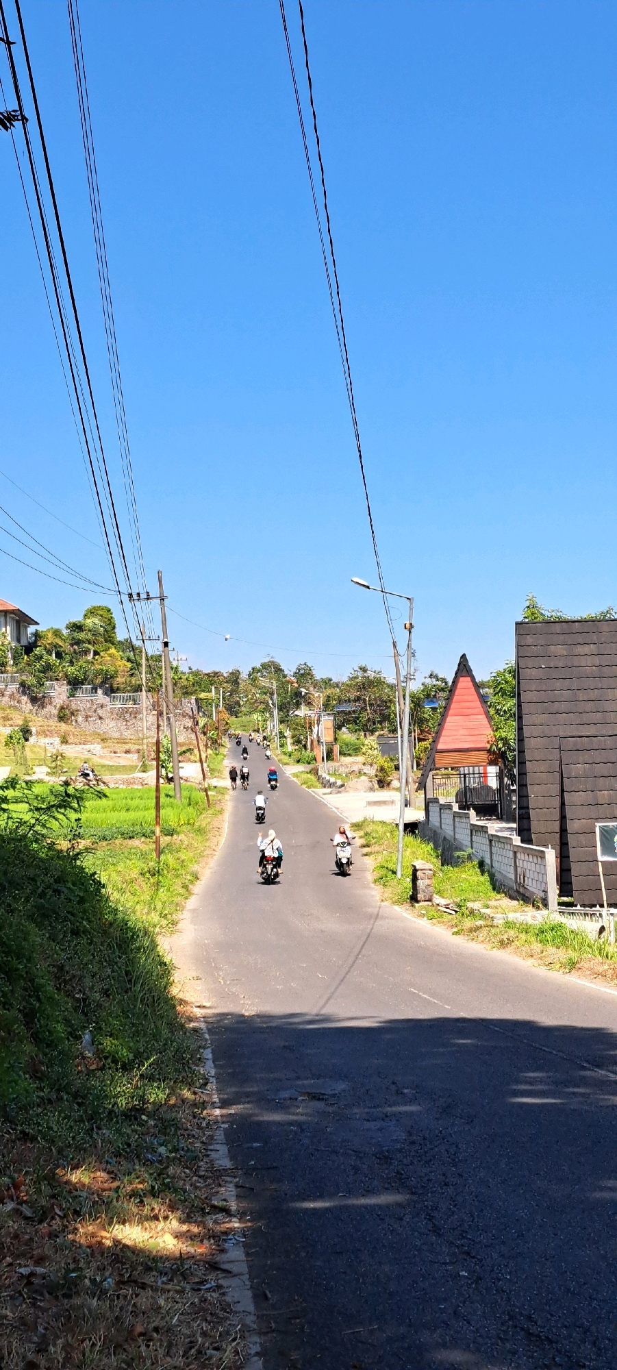 The image depicts a rural road in Kesiman, Indonesia, on a clear, sunny day. The road is paved and appears to be in fair condition, with some visible patches. A procession of motorcycles is traveling uphill, with at least seven visible in various positions on the road. On the left side of the road, a steep, vegetated embankment slopes upwards, covered in dense green grass and some dry brush at the bottom. Further up the embankment, there are signs of human habitation with stone walls and buildings. On the right side of the road, there are several structures. A distinctive red-and-orange triangular building with wooden slats is prominent, and next to it is a dark-tiled roof structure. A white brick wall separates the road from these buildings. Overhead, a network of power lines crisscrosses the sky, supported by utility poles. The sky is a vibrant, cloudless blue. The overall scene suggests a typical day in a rural or semi-rural area of Indonesia, with motor vehicles being a primary mode of transportation.