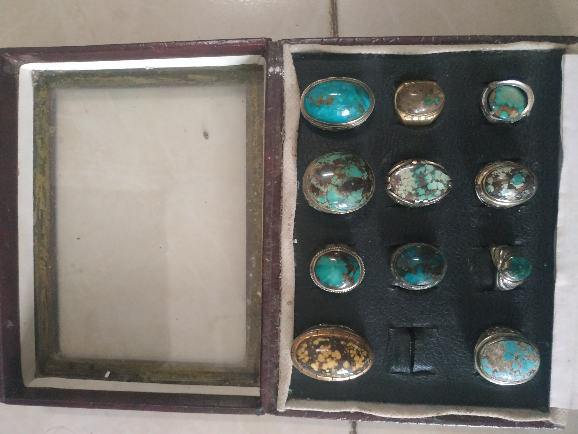An open, dark brown, leather-lined jewelry box with a glass lid is displayed on a light-colored, tiled surface. The box contains twelve rings arranged in four rows. The top row has three rings, the second and third rows each have four rings, and the bottom row has one ring visible. The rings feature various colored stones, predominantly turquoise and brown hues, set in metal bands. The environment is indoors. This image was taken in the city of Arjasa, Indonesia.