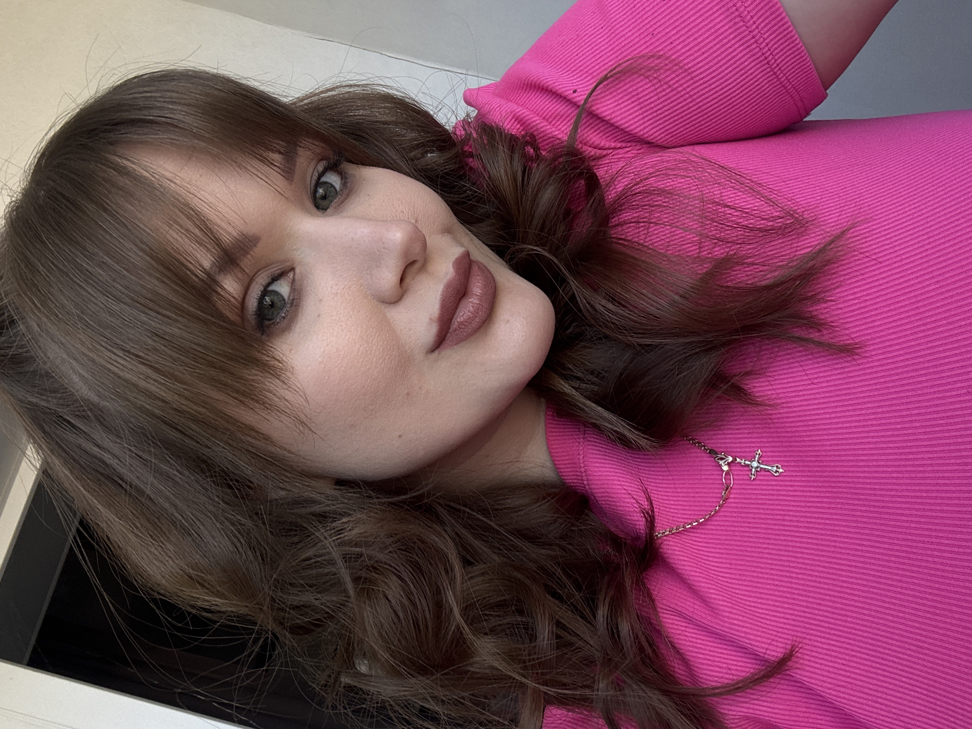 A woman with brown hair and bangs is pictured in a close-up selfie. She is wearing a bright pink ribbed top and a silver necklace with a cross pendant. The background appears to be a wall, possibly indoors. There is no discernible information in the image that would indicate the specific location in Chaikovsky, Russia.