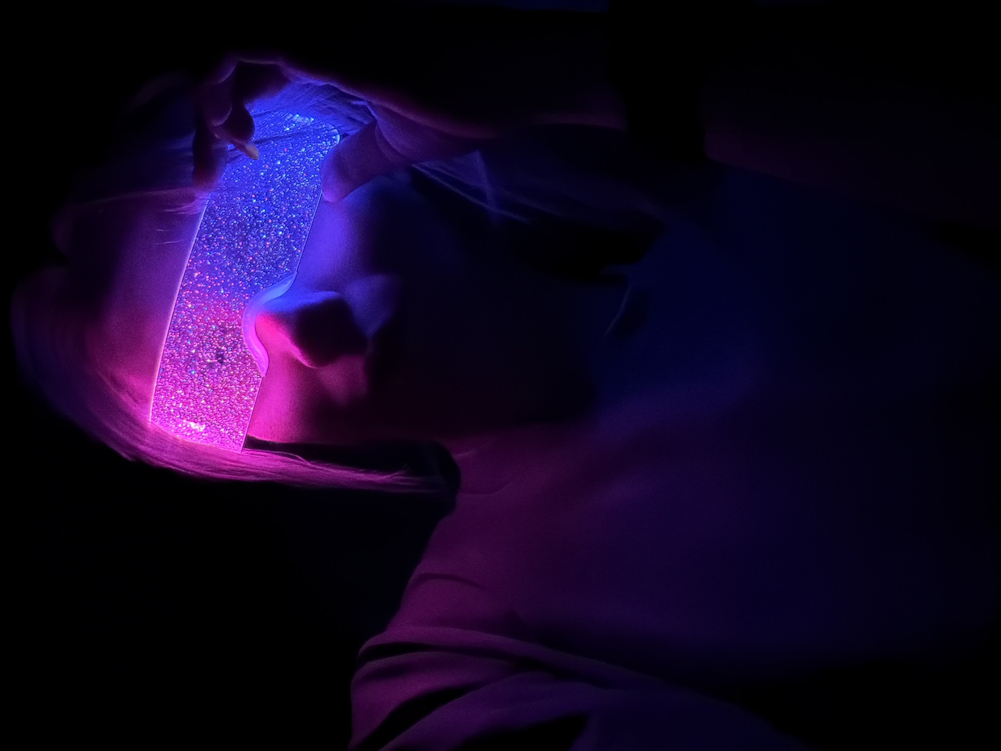 A person's silhouette is illuminated by a glowing rectangular object with glitter inside, held near their face in a dark environment. The light casts blue hues on the upper portion and pink hues on the lower portion of the object. The person's face is obscured by the shadow, but the nose and part of the cheek are visible. The lighting also casts shadows on the person's neck and shoulder. The surrounding environment is black, suggesting a dimly lit or nighttime setting. The location in Chaikovsky, Russia is not discernible from the image.