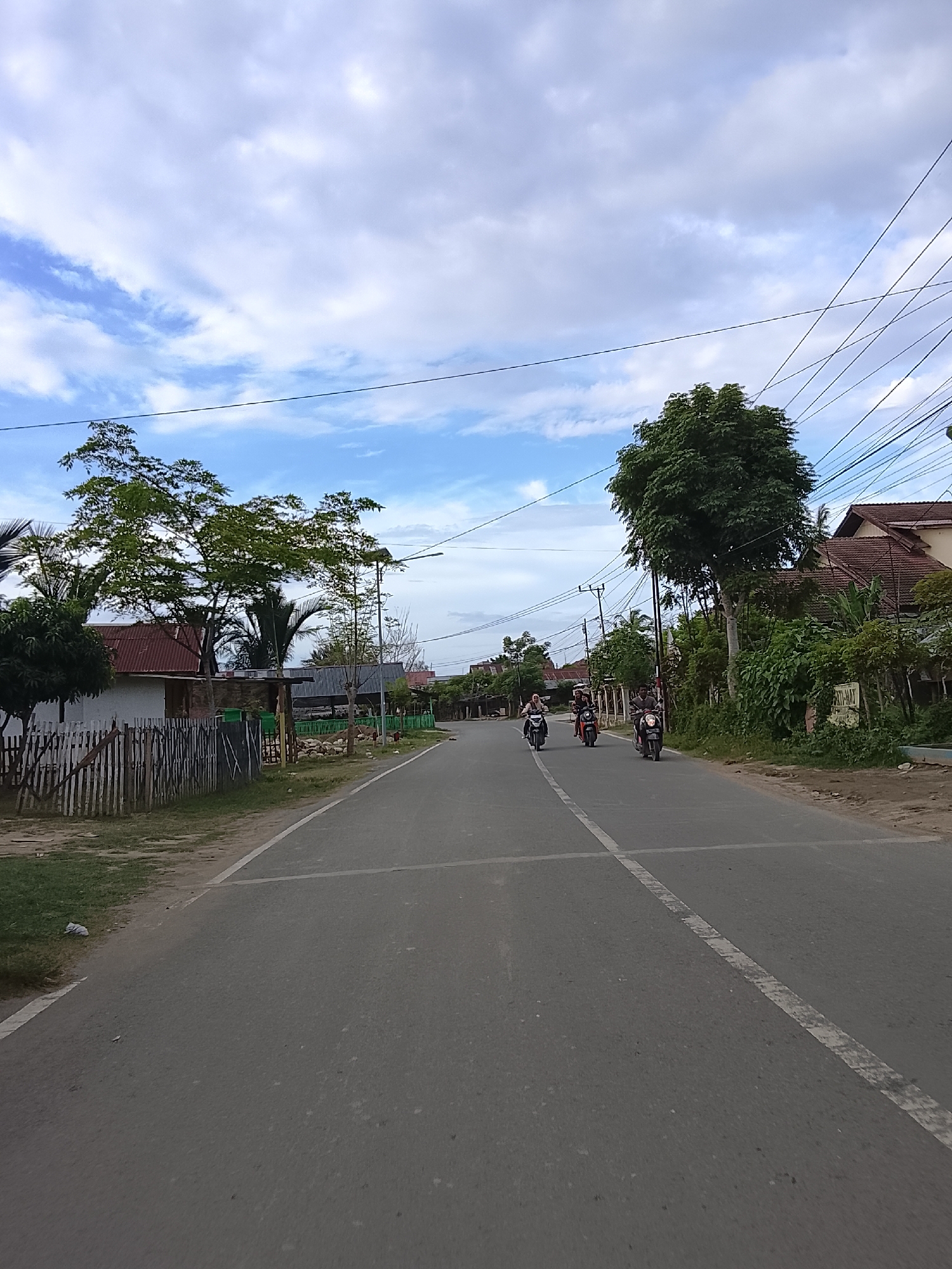 The image shows a straight, paved road in Blang Cut, Indonesia, under a cloudy sky. Several motorcycles are visible on the road, with riders on each. On the left side of the road, there is a wooden fence, some grass, and houses with red roofs. On the right side of the road, there are trees, more houses, and utility poles with numerous wires. The scene depicts a typical day on a suburban road in Indonesia, with ongoing motorcycle traffic and residential infrastructure.