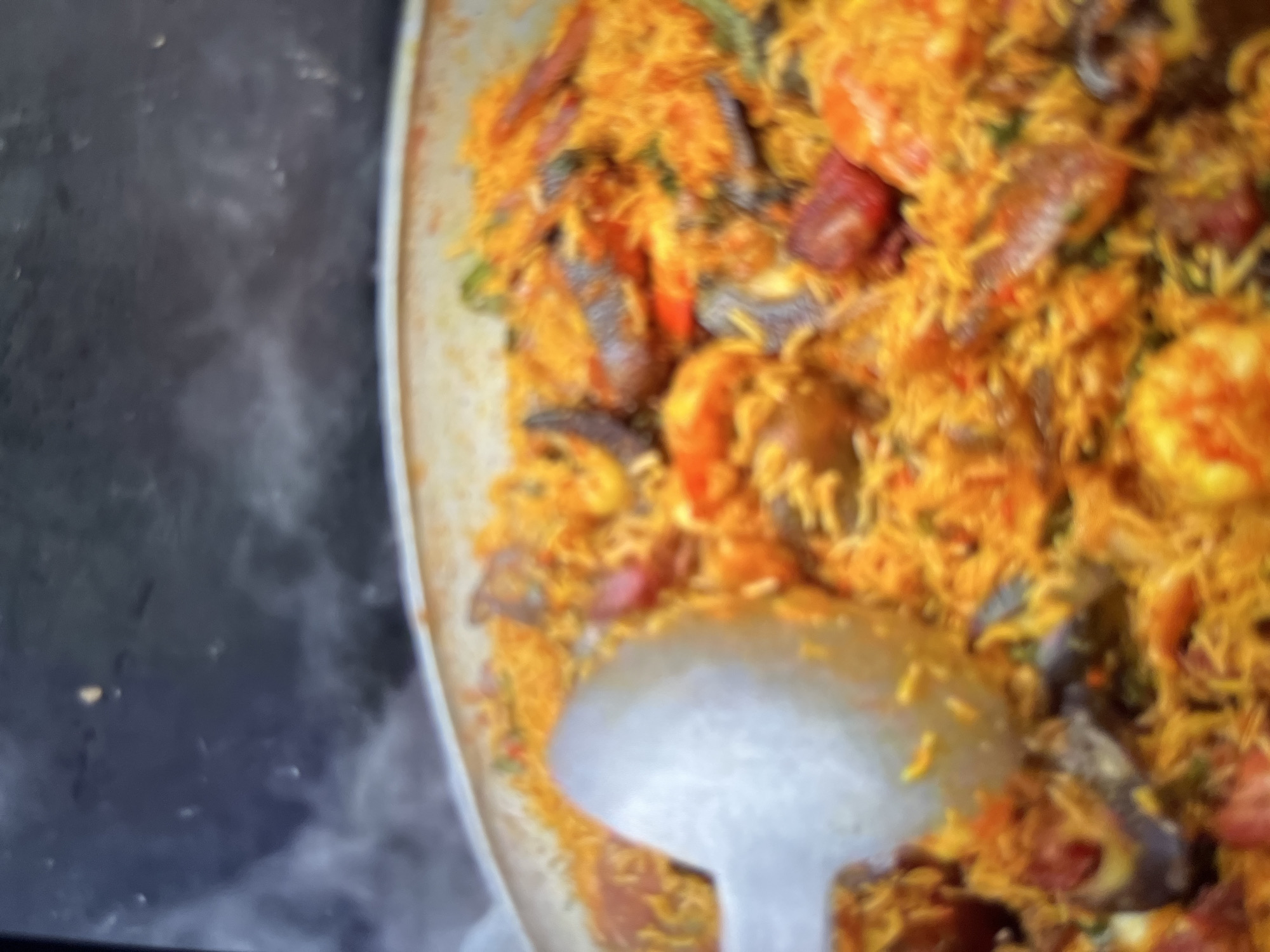 The image shows a close-up of a large pan filled with paella, a Spanish rice dish. The paella appears to contain rice, shrimp, mussels, and possibly chorizo sausage, all cooked in a rich, orange-hued broth. A large metal serving spoon is visible on the left side of the pan, partially submerged in the dish. Wisps of steam rise from the paella, indicating it is hot and freshly prepared. The background is dark and out of focus, but appears to be a kitchen or serving area. There are no people or specific landmarks of Abuja, Nigeria, visible in the image.
