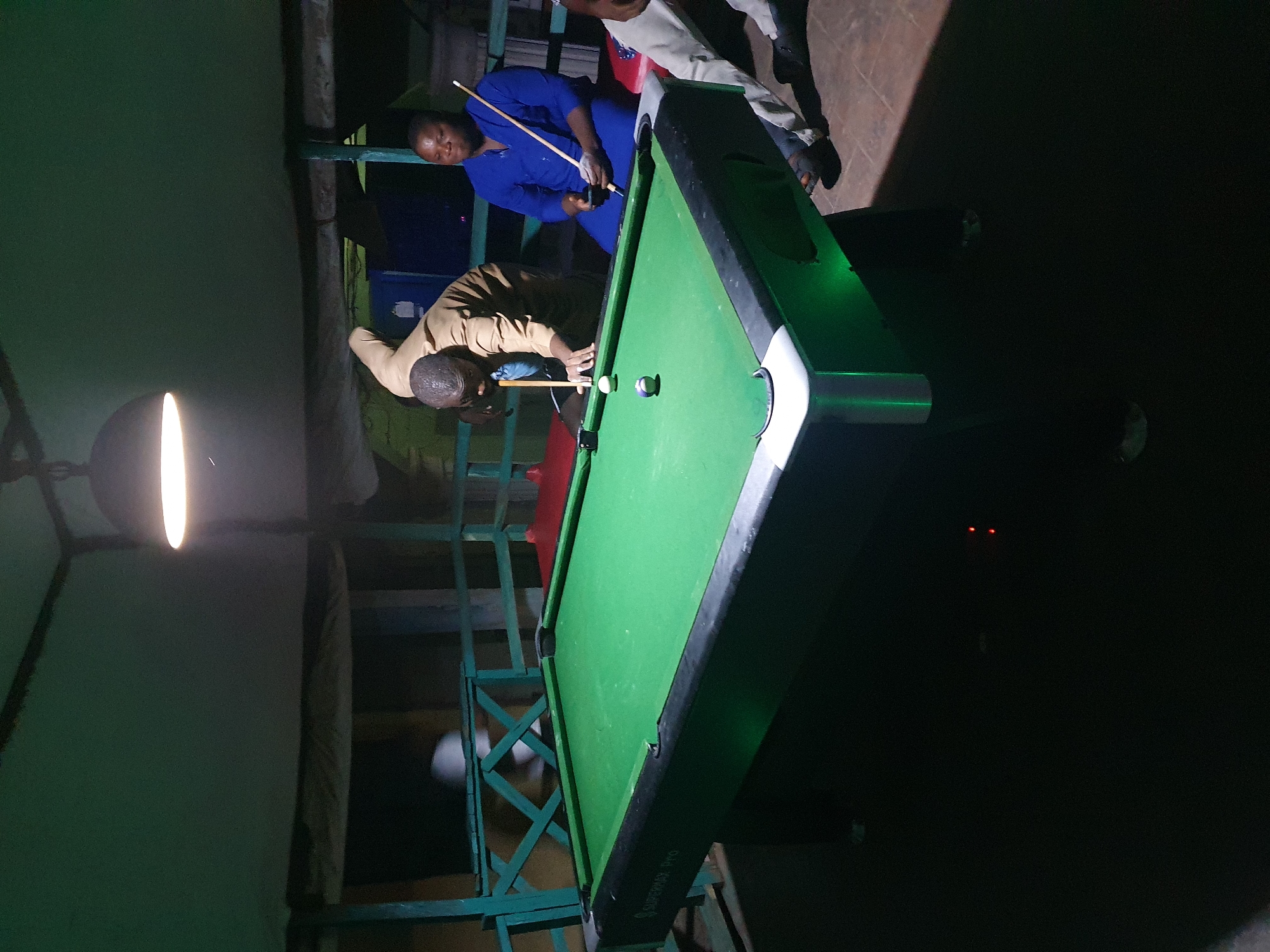 The media file shows a dimly lit scene, possibly at dusk or night, with a pool table as the central object. Two individuals are present, engaged in playing pool. One person, wearing a blue outfit, stands by the table, holding a pool cue, seemingly waiting for their turn or observing. The other person, dressed in a light brown top and dark pants, is in the process of taking a shot, leaning over the green felt of the pool table. A cue ball and another ball are visible on the table. The setting appears to be an outdoor or semi-outdoor area, with visible metal structures supporting a roof or a platform, suggestive of a recreational space. A bright overhead light illuminates the pool table. In the background, a dark, shadowy area suggests the surrounding environment, with two small red lights visible in the distance. The overall atmosphere is casual and focused on the game. The image is oriented vertically. Text is visible on the side of the pool table, but it is partially obscured and illegible. The scene suggests a leisure activity taking place in Lokoja, Nigeria.