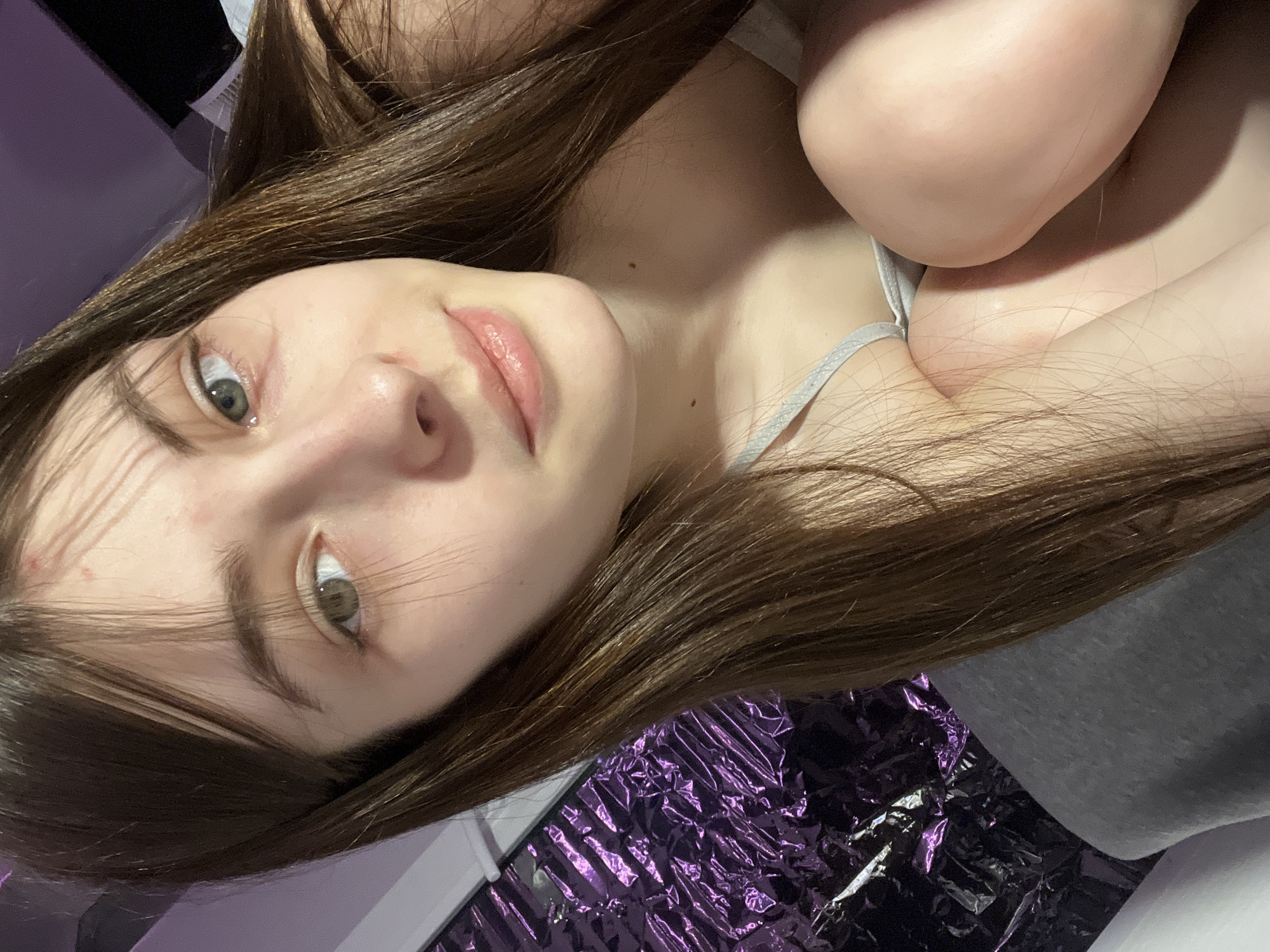 A close-up, angled selfie shows a young woman with long, brown hair looking at the camera. She has light skin, fair eyebrows, and light eyes. She is wearing a light-colored tank top. The background includes a purple, reflective material, possibly a sheet or wrapping paper. The location is not discernible from the image and there is no basis to suggest Saint Petersburg, Russia.